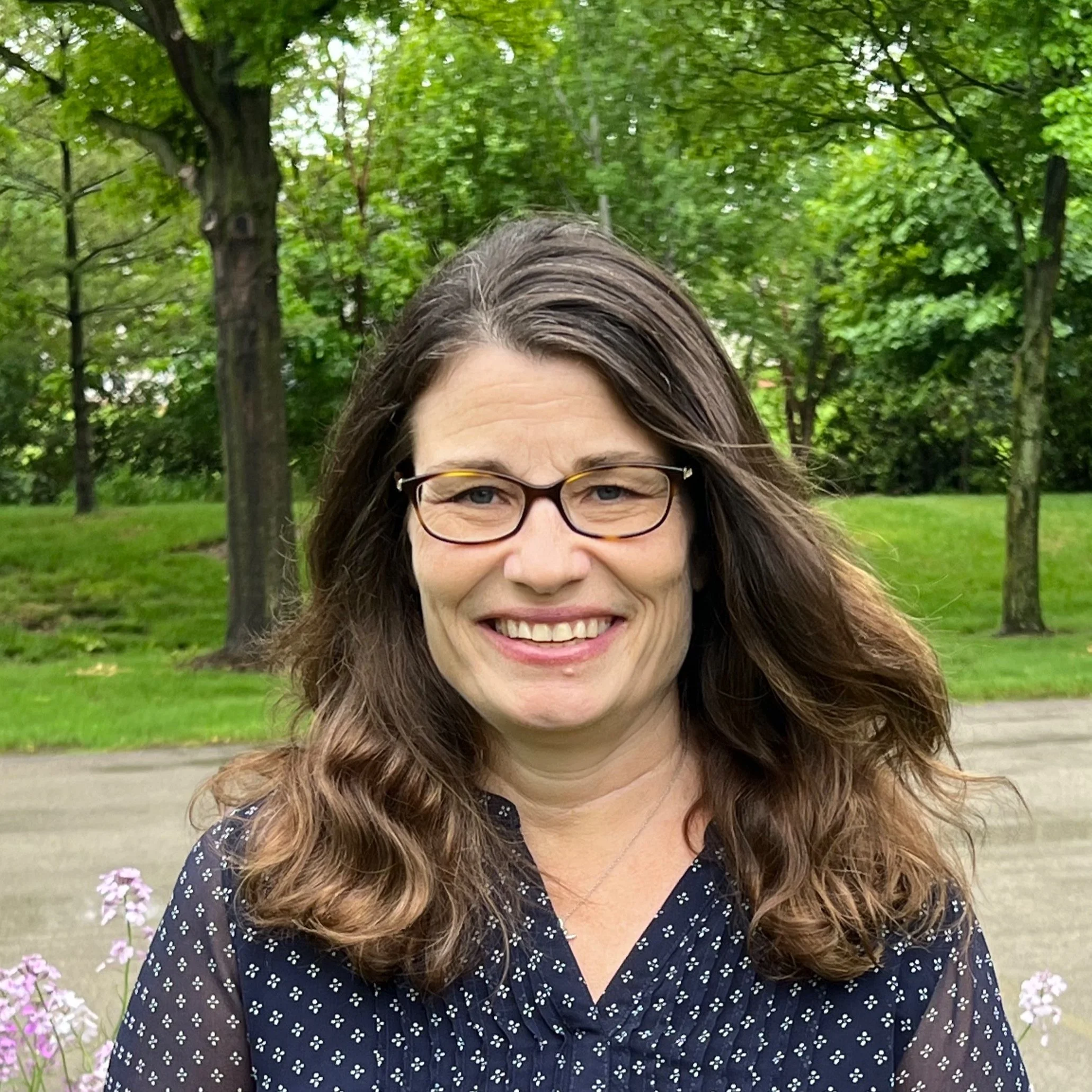 A woman with long wavy brown hair, wearing glasses and a navy blue shirt with white polka dots, smiling outdoors in a park with green trees and grass in the background.