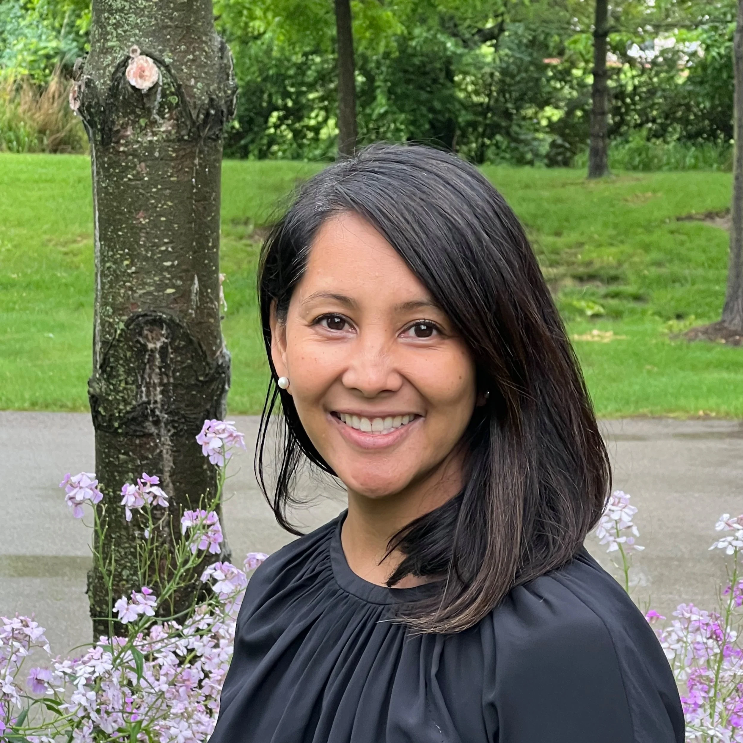 A woman with shoulder-length black hair smiling outdoors near a tree with purple flowers, green grass, and other trees in the background.