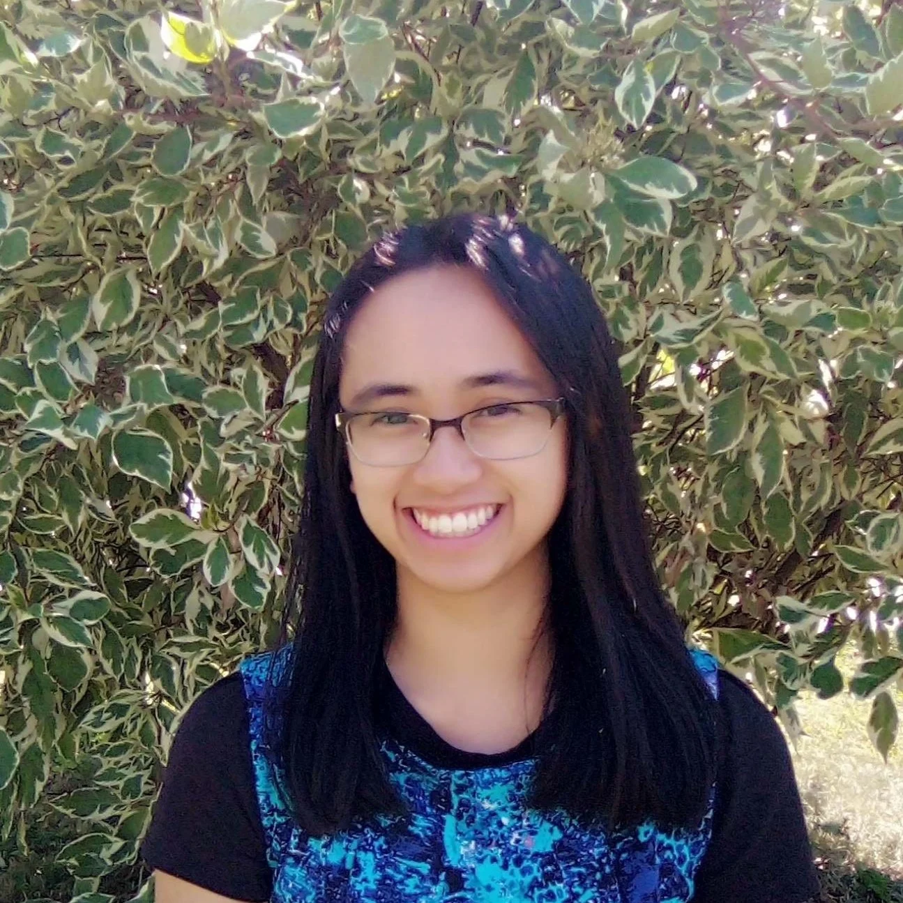 Young woman with glasses and long black hair smiling outside in front of a bush with variegated green and cream leaves.