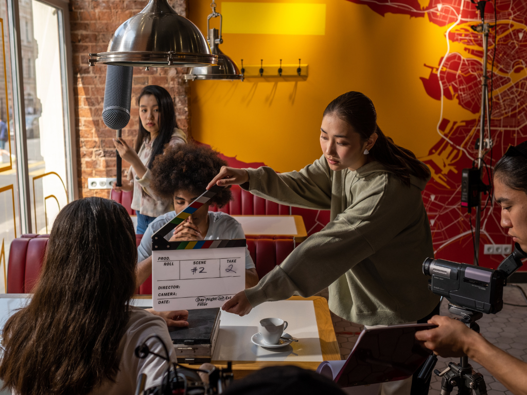 Filmmaking crew on set with a clapperboard, camera, and microphone in a cozy restaurant or cafe with red booths and yellow walls.
