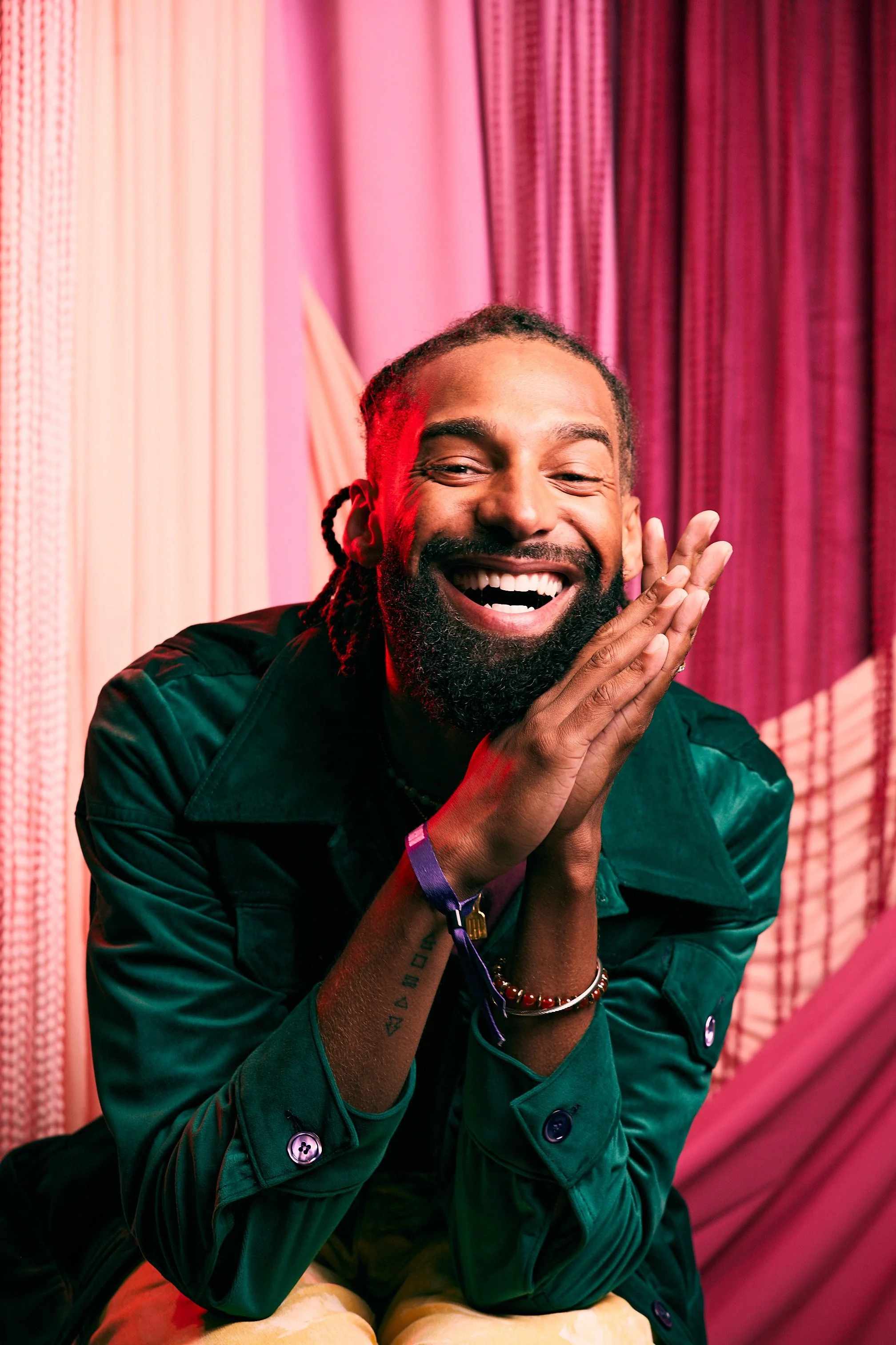 A man with a beard and dreadlocks smiling with his eyes closed, wearing a dark green jacket, sitting in front of pink and purple curtains.
