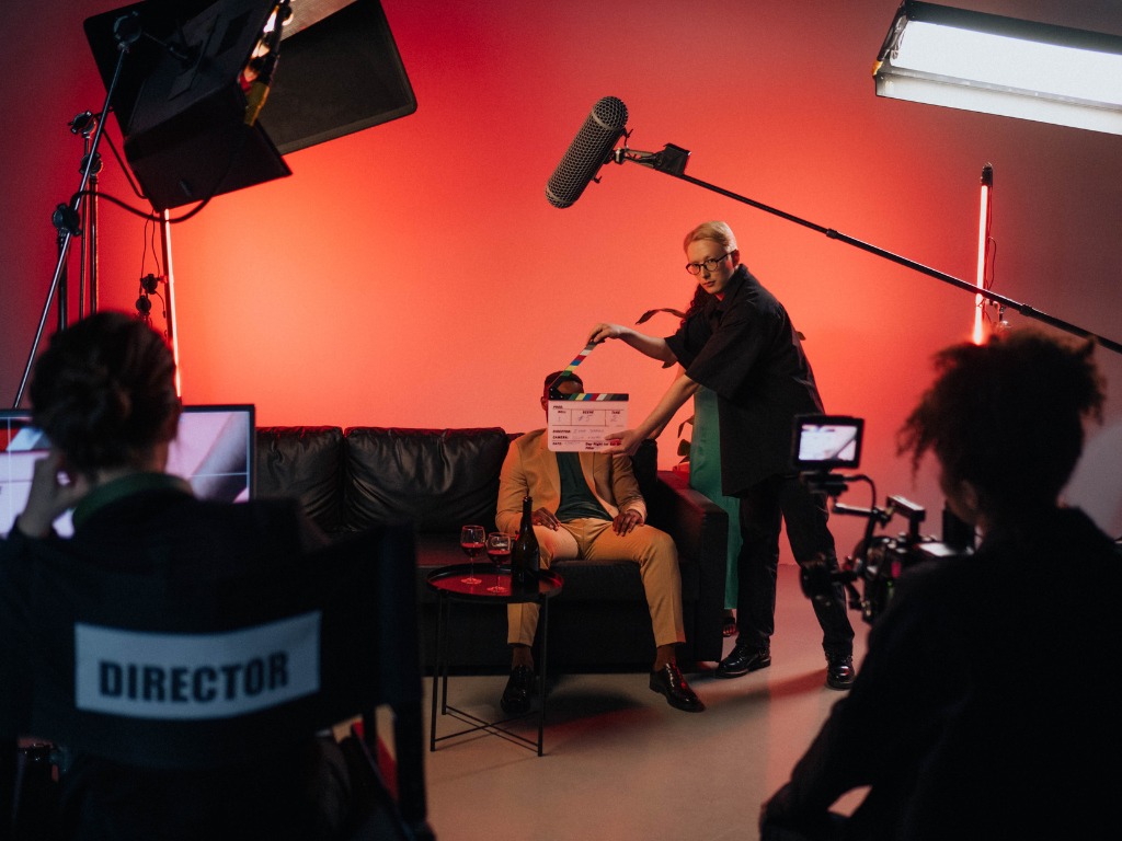 Film crew on set with a woman holding a clapperboard, a person sitting on a black couch with wine glasses and a bottle, and a director's chair labeled 'Director'. The scene is lit with studio lights and a red backdrop.