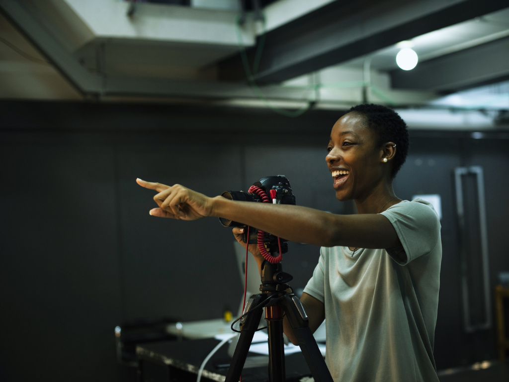 A woman with short curly hair smiling and pointing at something, holding a camera on a tripod in a studio or indoor space.