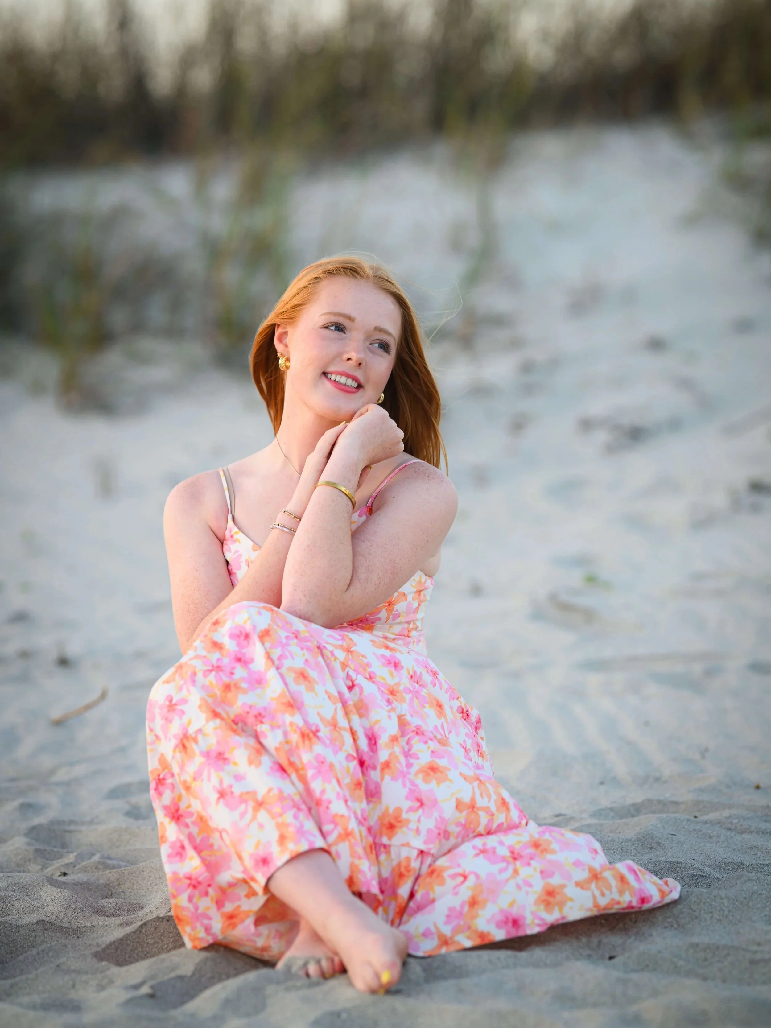 A woman standing ankle-deep in the ocean waves at sunset, wearing a pink sleeveless top and a long beige skirt with floral embroidery, looking at the camera with a slight smile.
