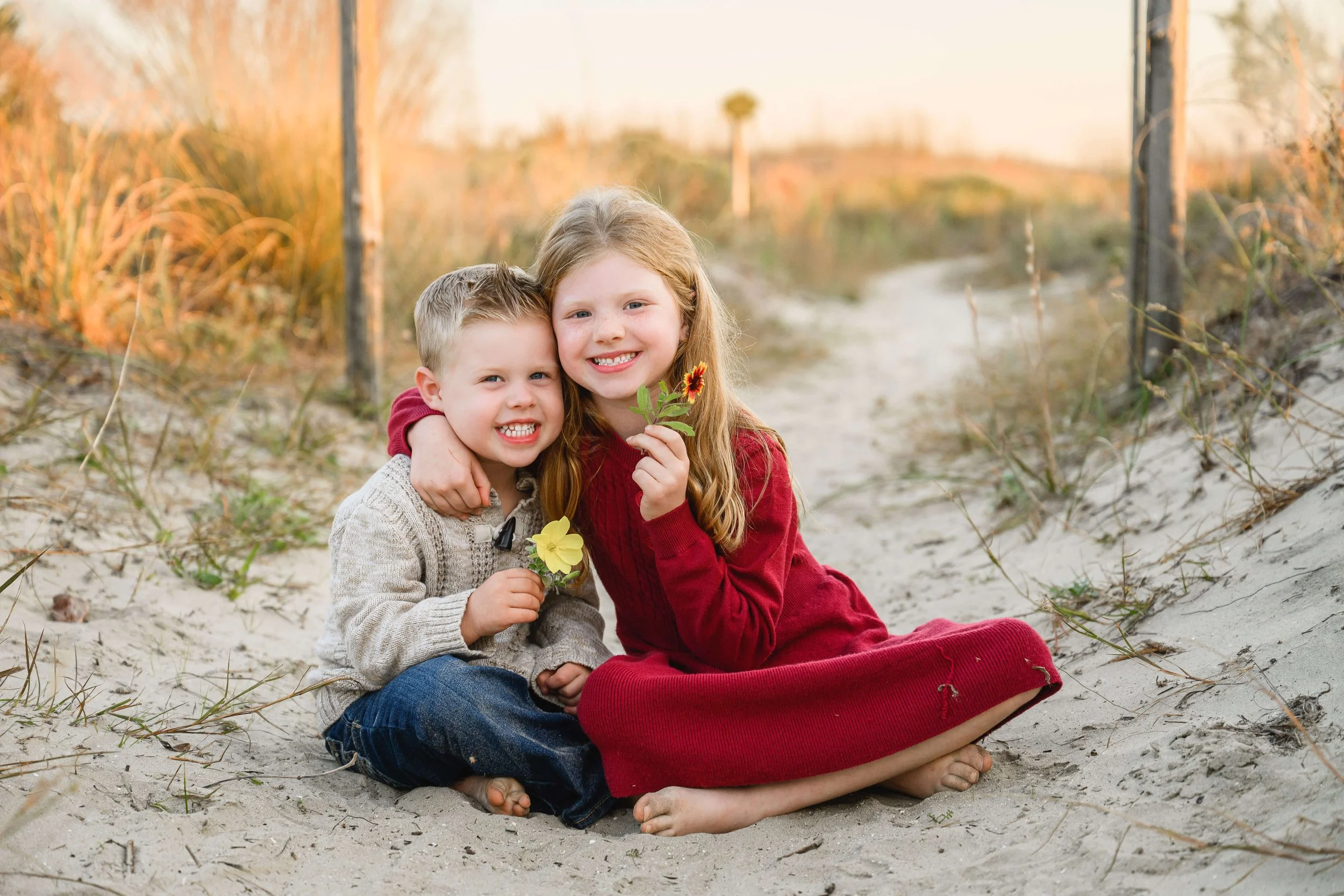 Two siblings sitting together in the sand at Isle of Palms holding small yellow flowers.