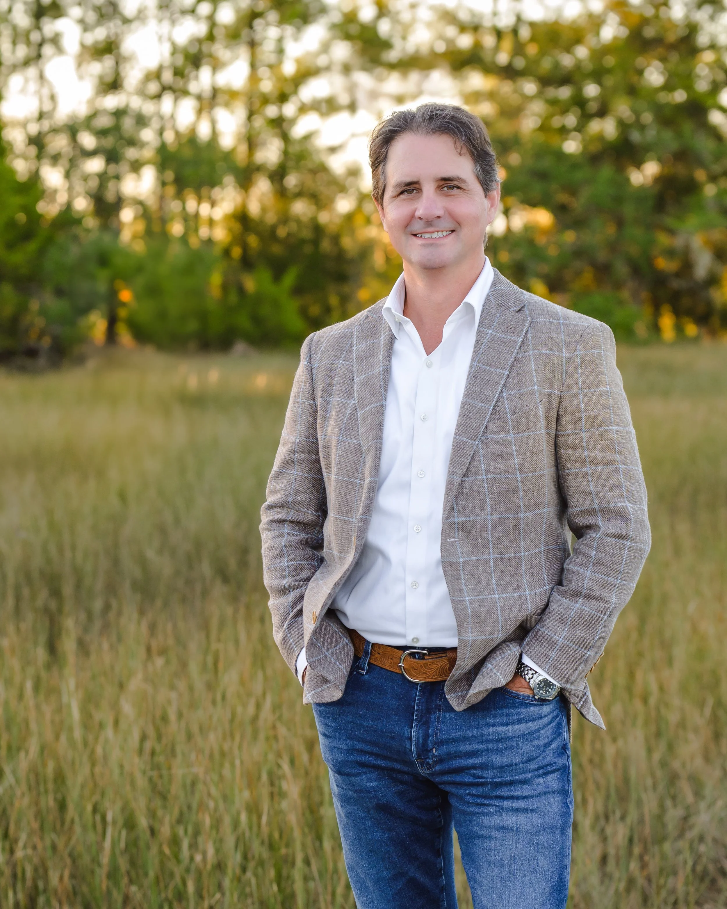 Man in maroon shirt and jeans leaning on a wooden railing on a dock by the water with trees and a bridge in the background.