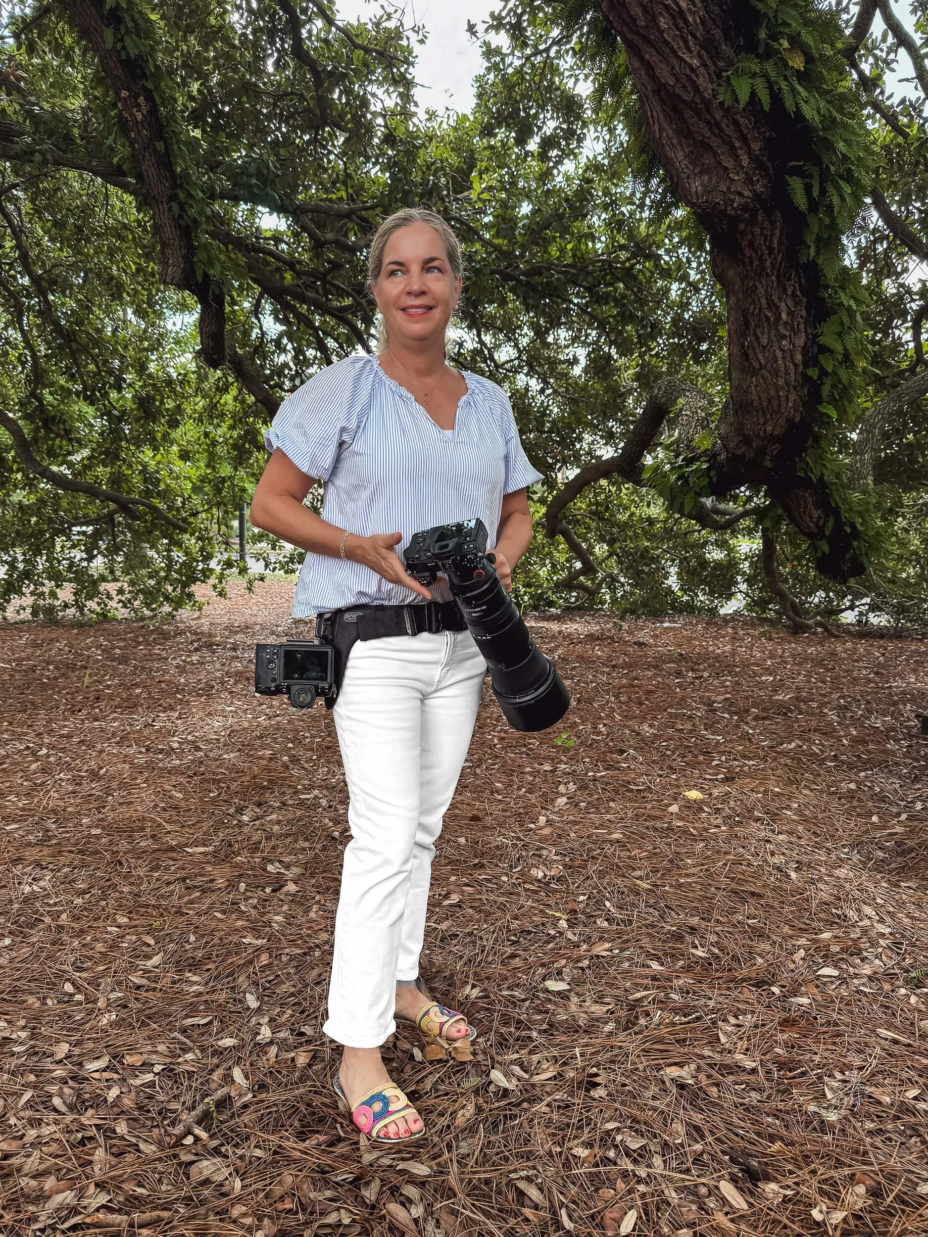 A woman standing outdoors on a dirt ground with pine needles, holding a camera with a large telephoto lens, wearing a blue and white striped blouse, white pants, and colorful sandals, in front of a large tree with lush green foliage.