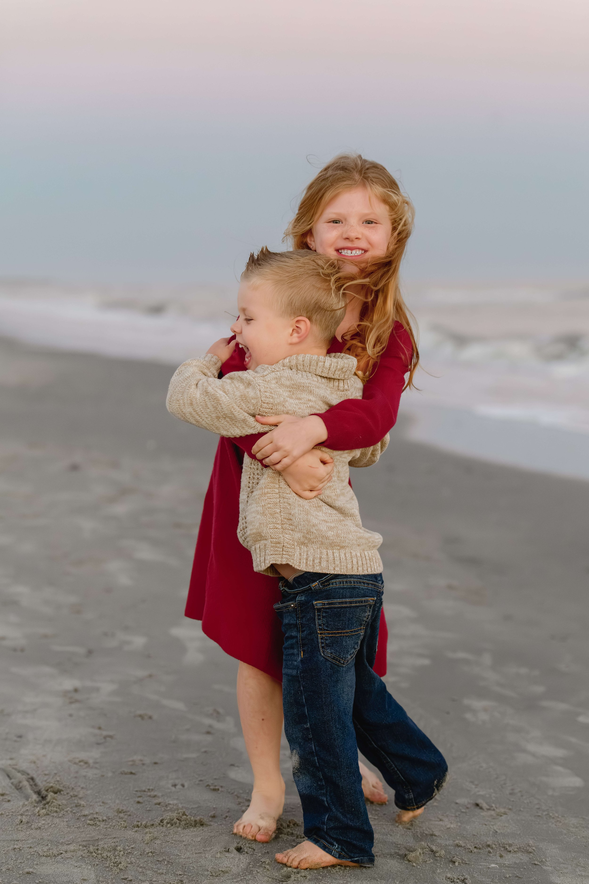 Older sister in a red dress hugging her younger brother on the sand at Isle of Palms.