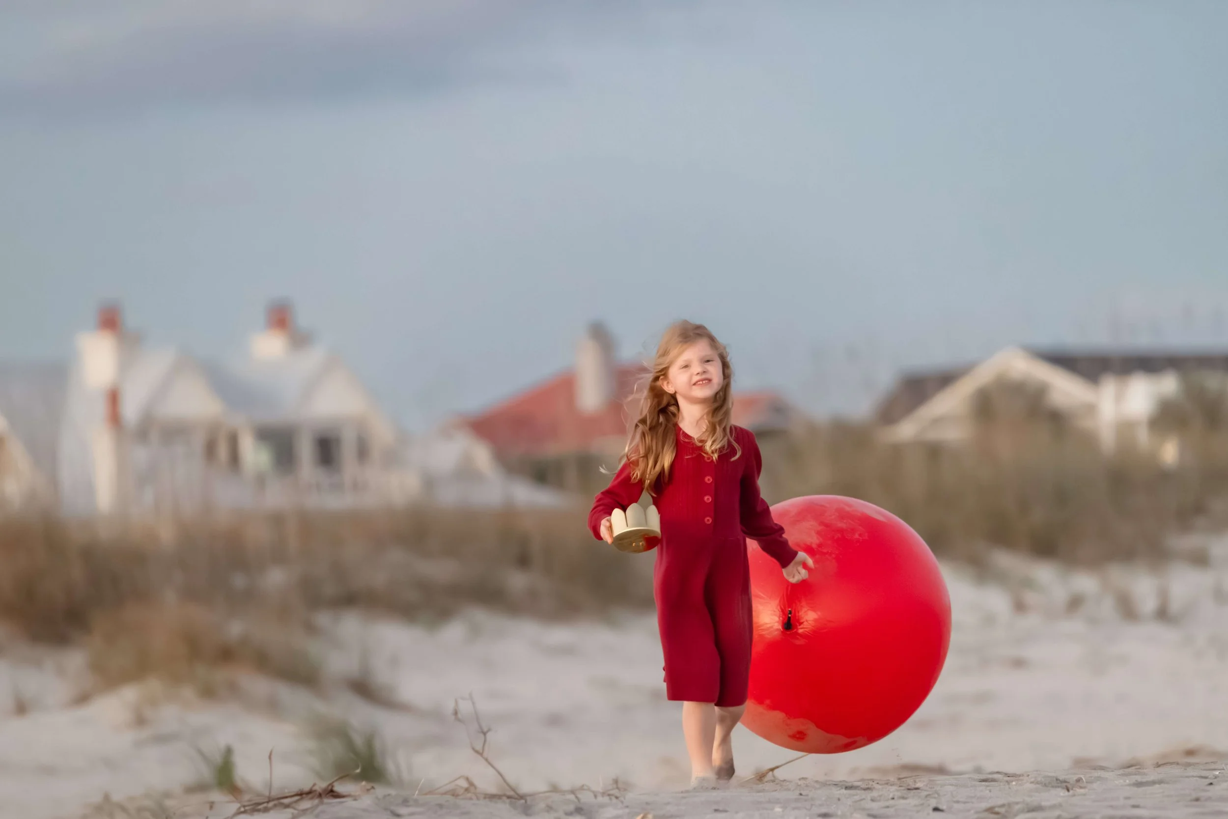 A young girl running through the sand holding a large, shiny red Christmas ornament.