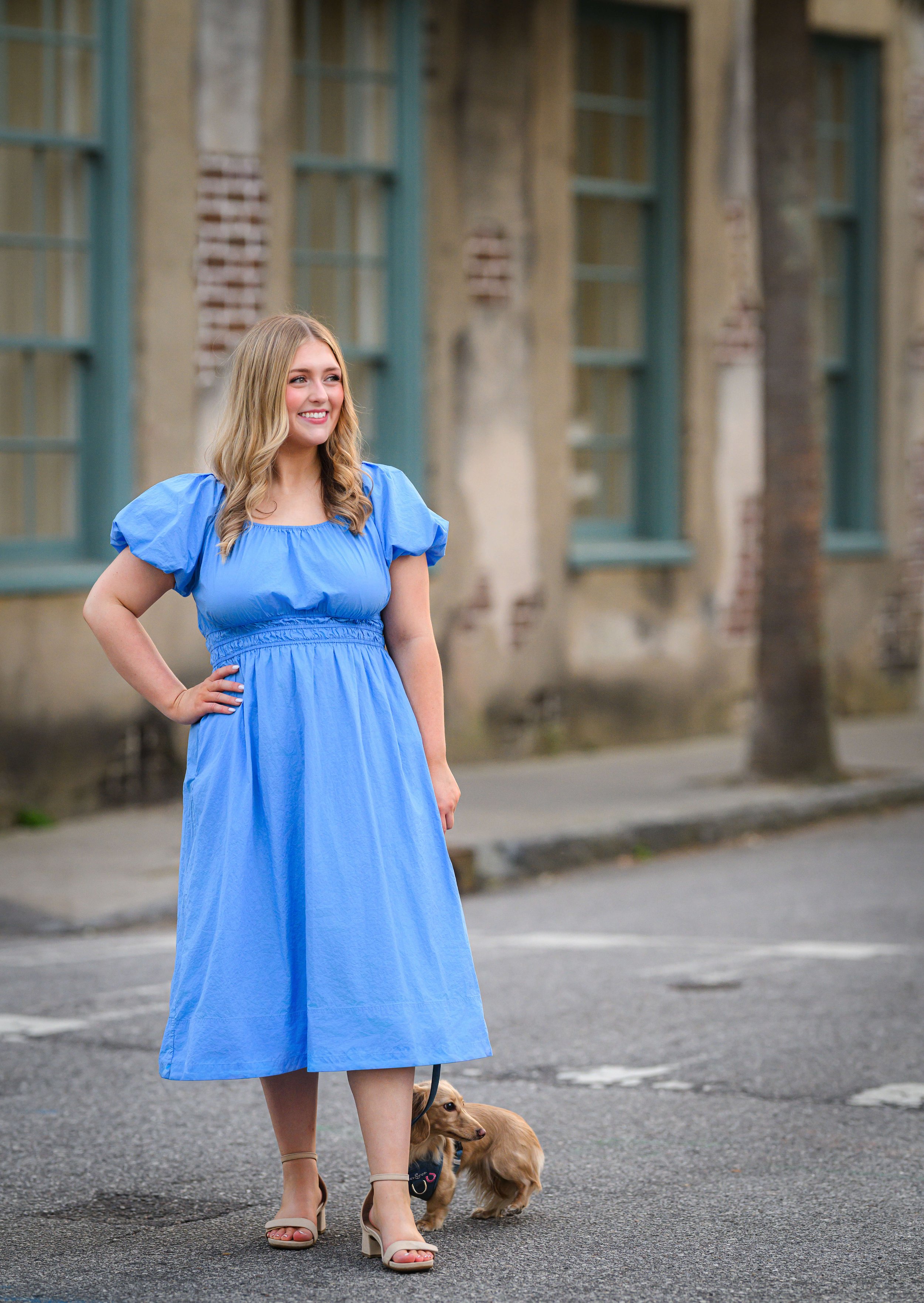 A woman in a blue dress walking a small brown dog on a Charleston street in the French Quarter with old brick buildings and green window frames in the background.