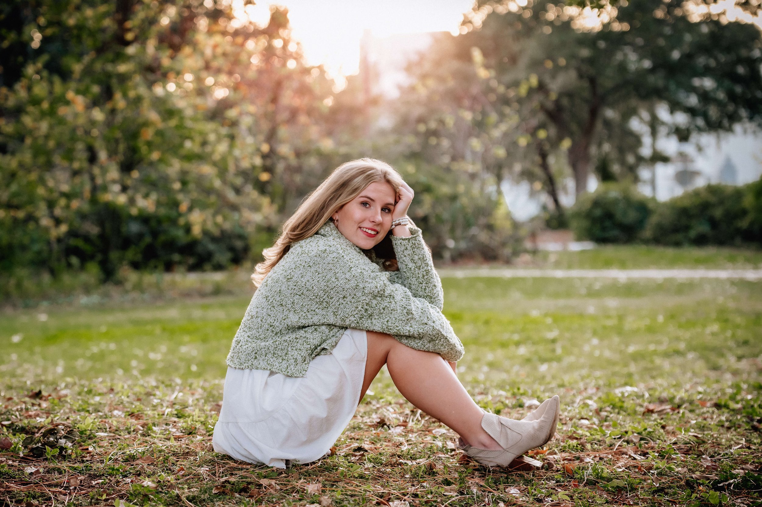 Girl wearing light green sweater over white dress with ankle boots sitting on a field in Hampton Park, Charleston, SC