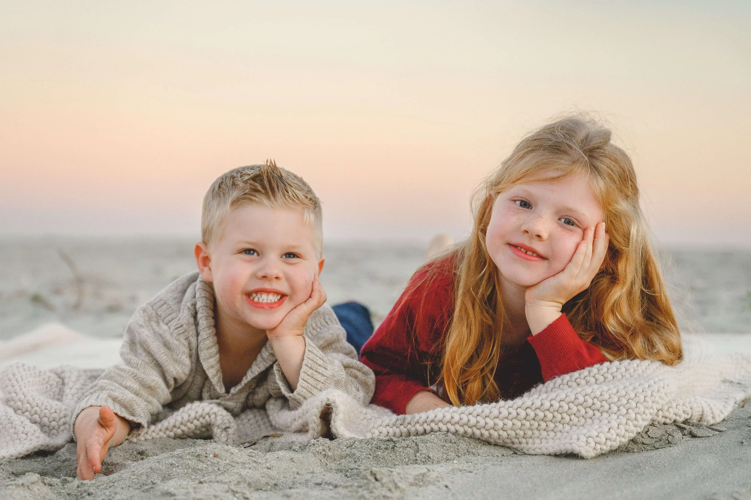 Two young siblings in fall sweaters smiling on a cream knit blanket at Isle of Palms beach.
