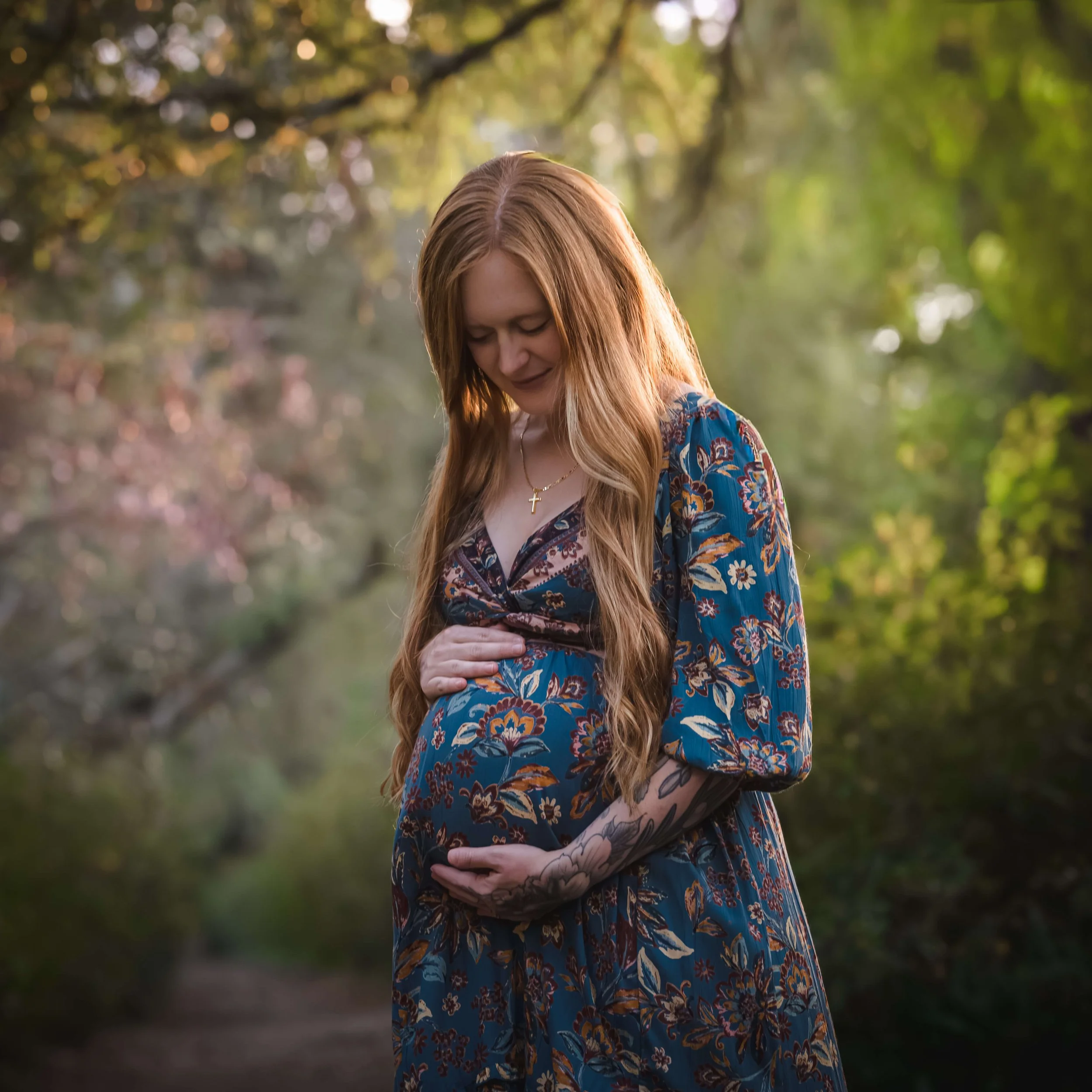A pregnant woman with long red hair, wearing a floral dress, is standing outdoors in a wooded area, gently touching her belly and looking down affectionately.