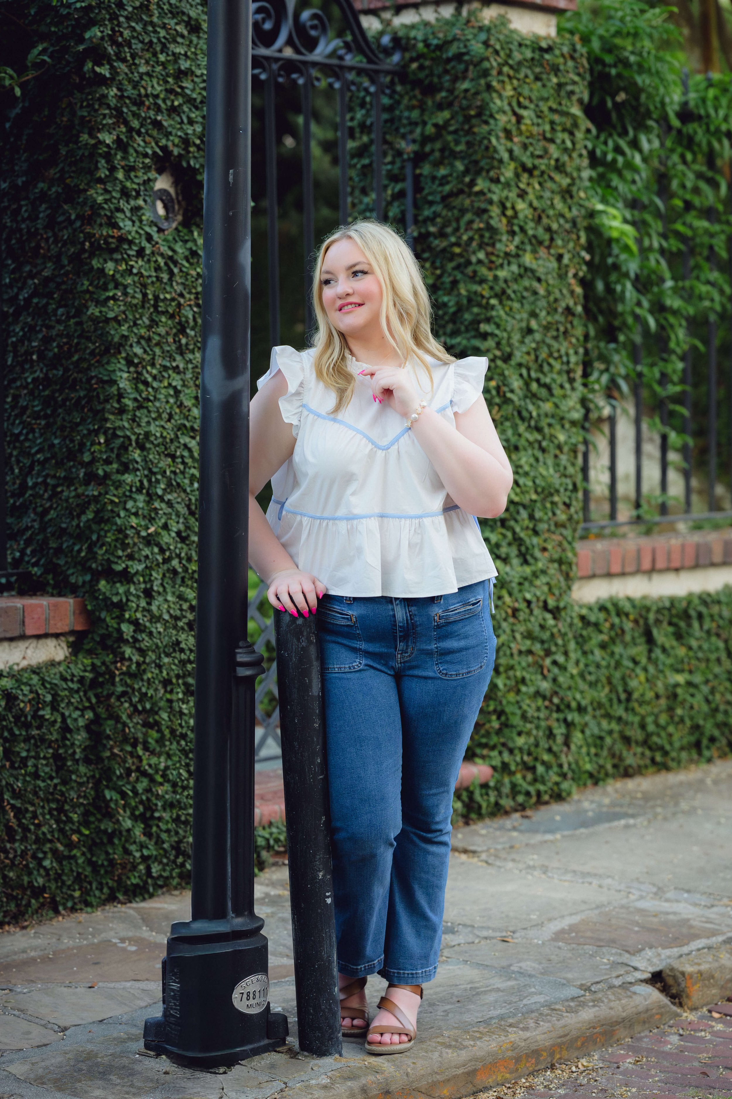 A young woman with blonde hair leaning on a black streetlamp pole in Charleston with greenery in the background, wearing a white ruffled top, blue jeans, and beige sandals.