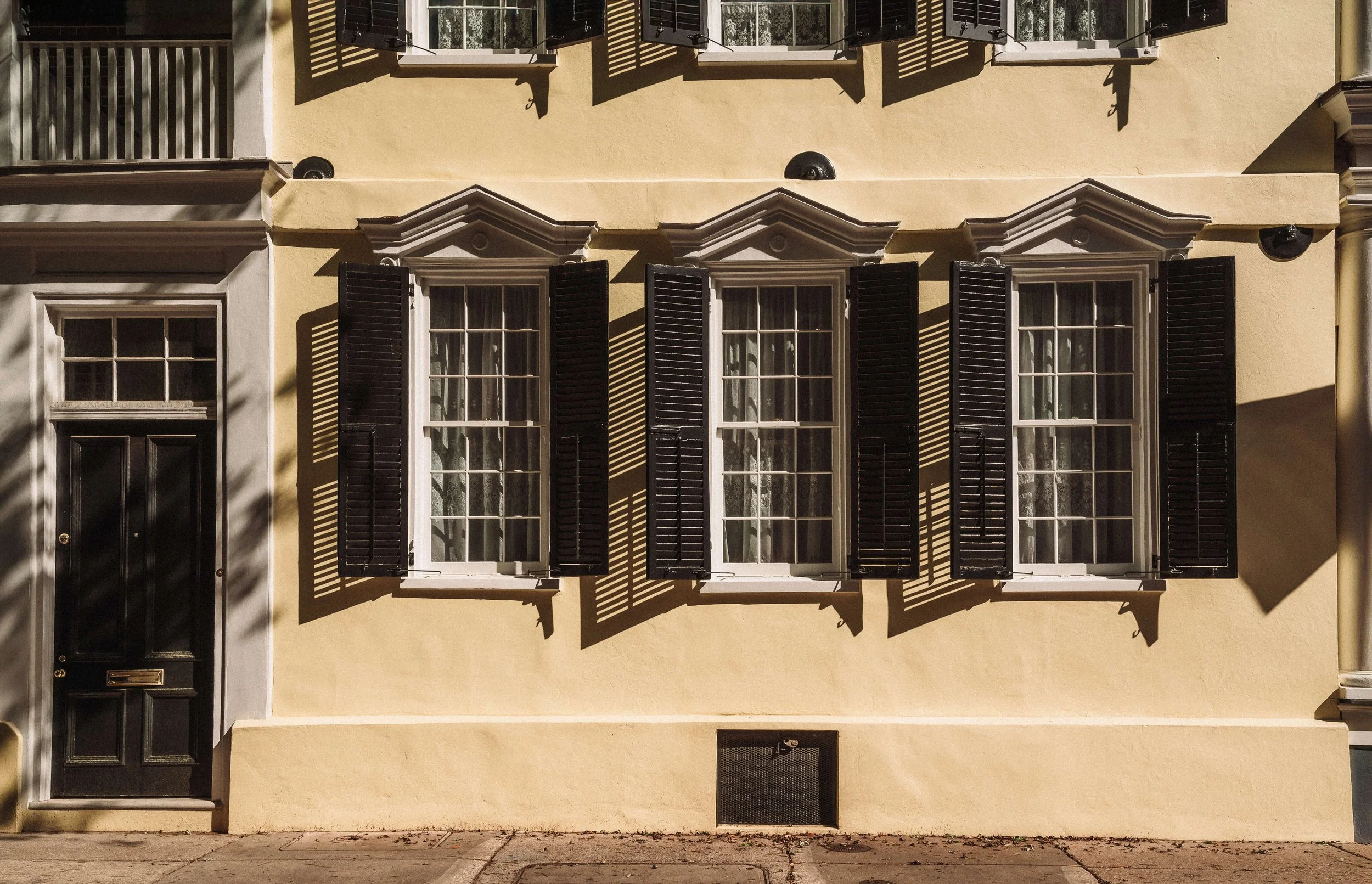 A yellow building with three large windows featuring black shutters, and a black door with a mail slot, all framed by white trim. Shadows cast by a nearby tree create a pattern across the facade in Charleston.