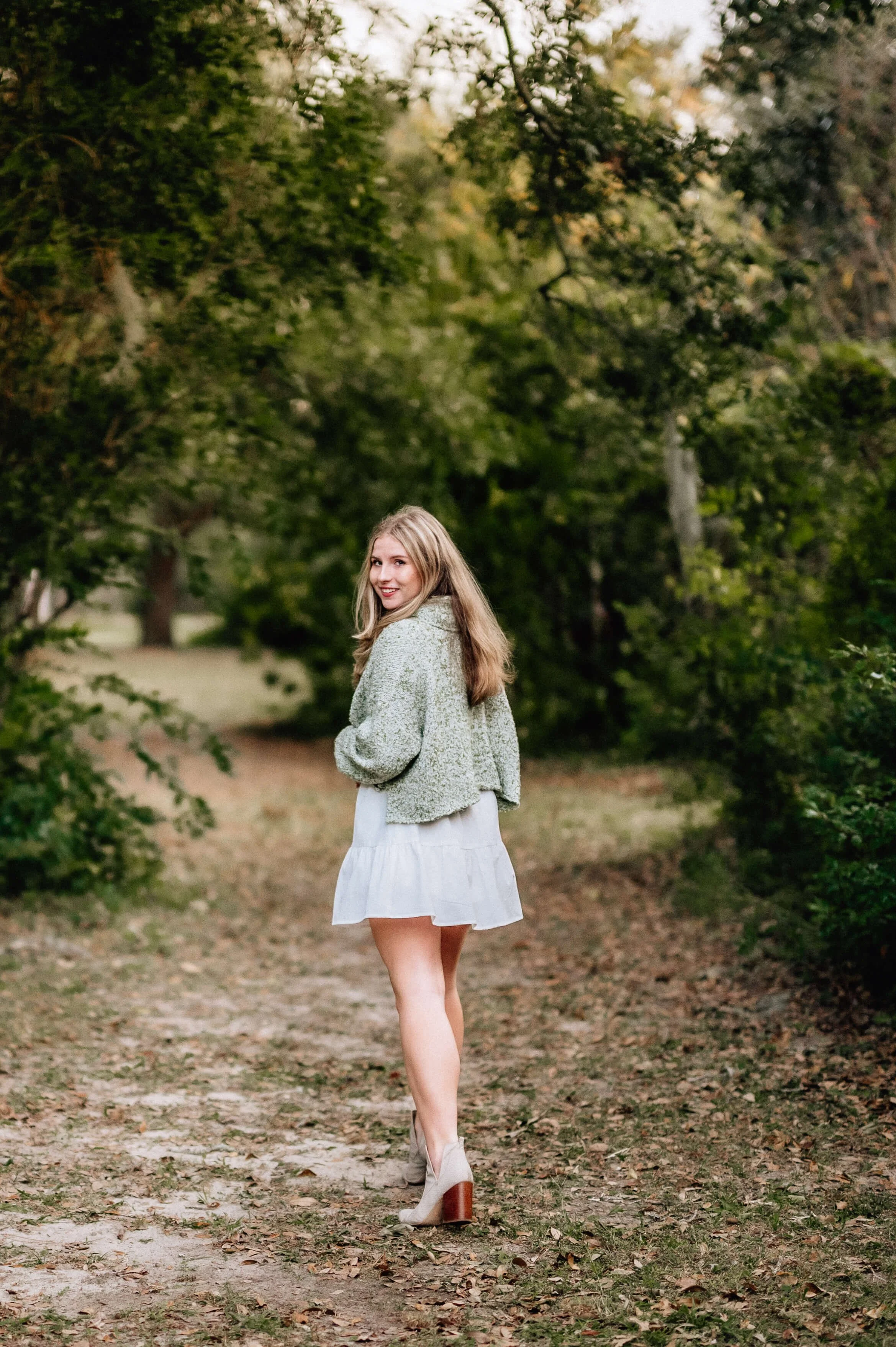young lady looking over her shoulder and smiling at the camera wearing a light green sweater and white skirt at Hampton Park in Charleston, SC