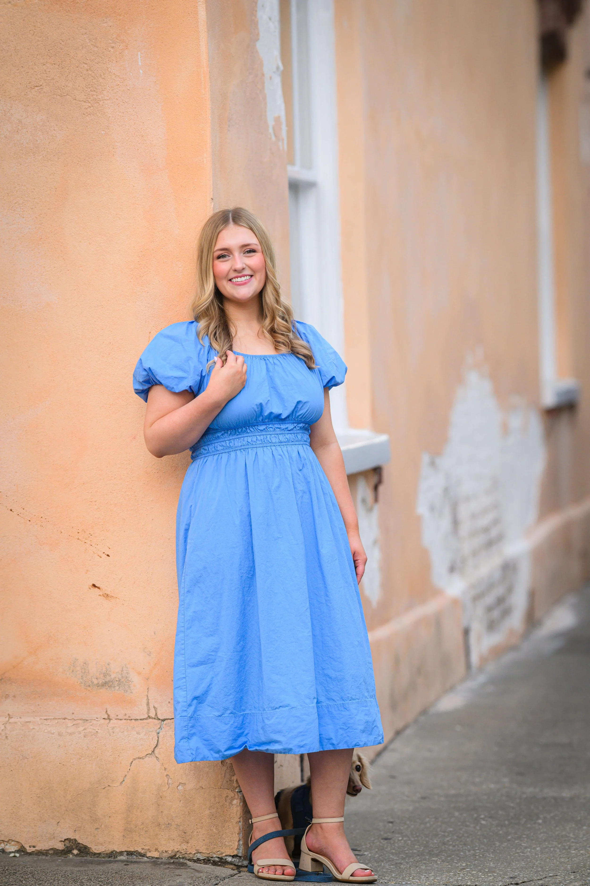 A young woman with blonde hair wearing a blue dress standing against a peach-colored wall in Charleston with some visible wear and cracks, smiling at the camera.