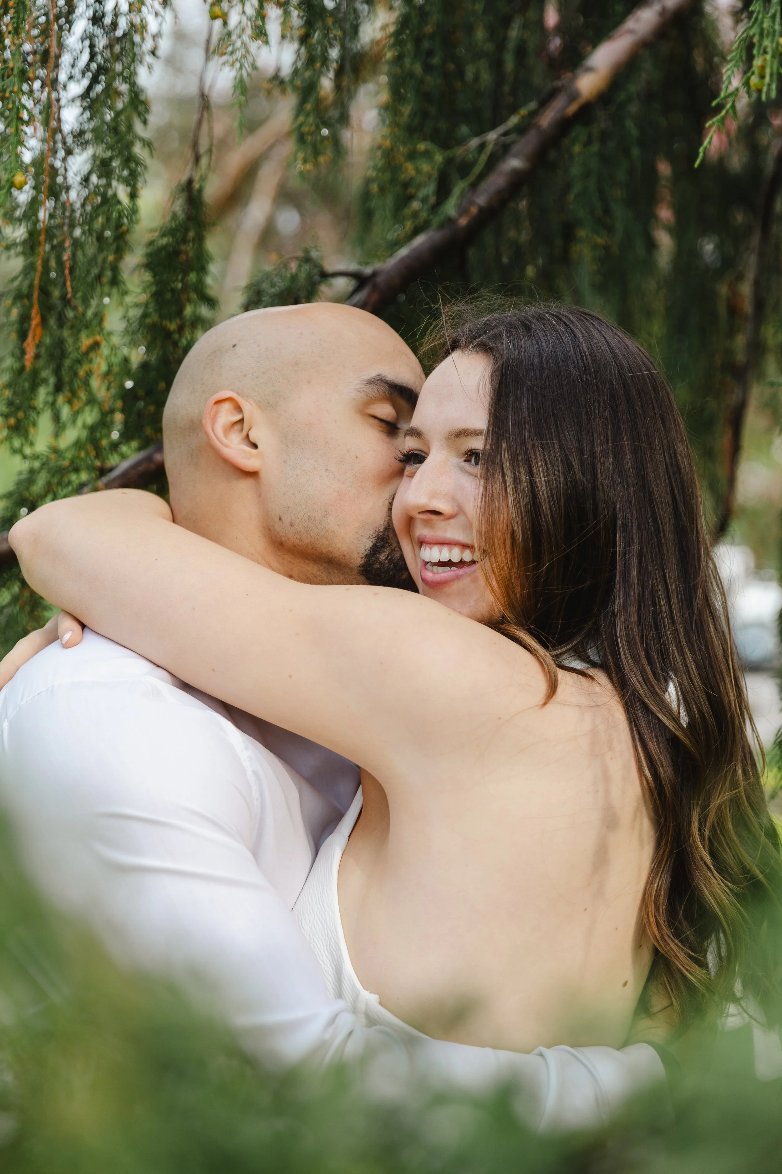 A couple is embracing outdoors among trees, with the man kissing the woman's forehead and both smiling.