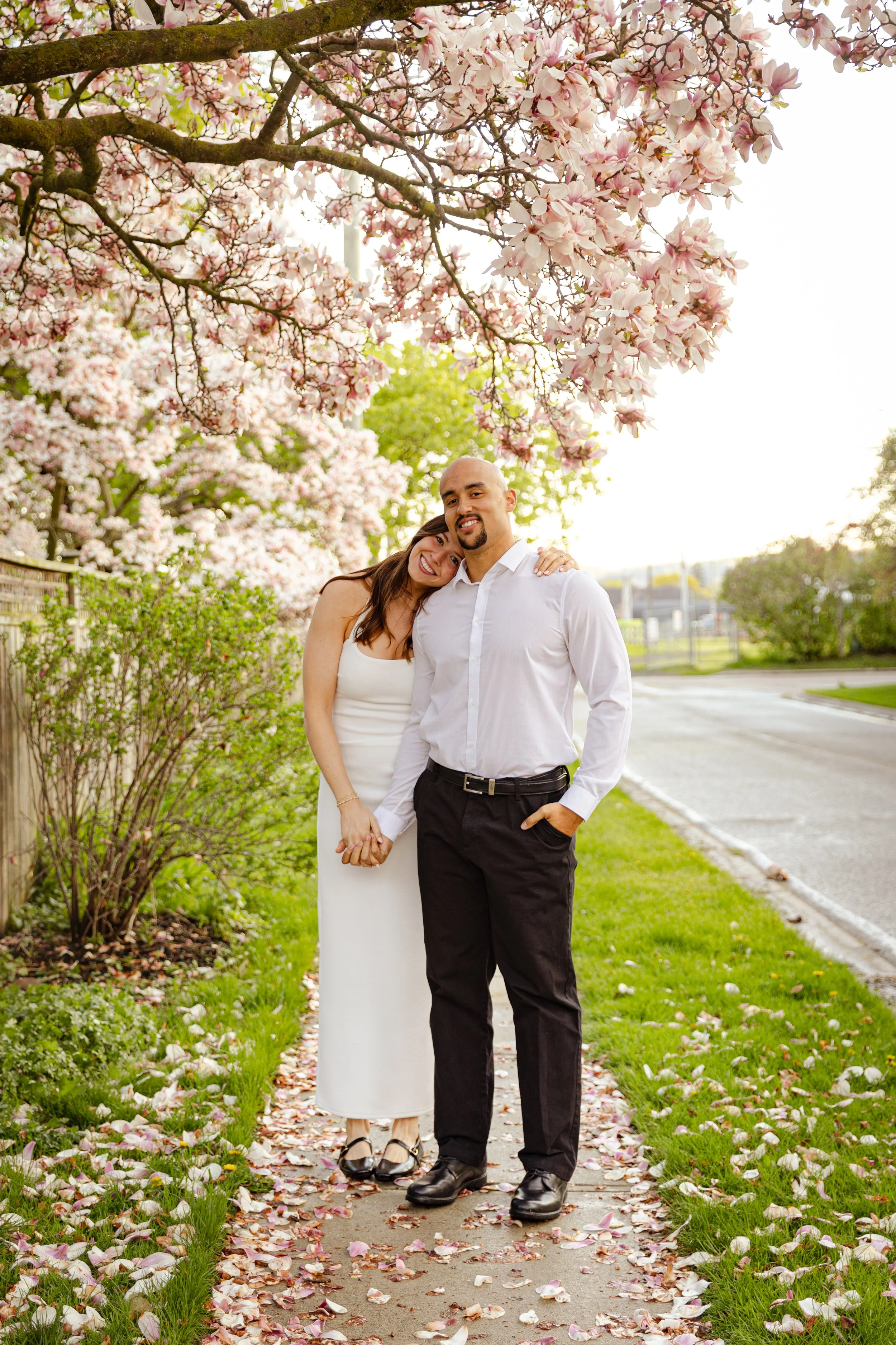 A couple standing on a sidewalk under a tree with pink and white blossoms, holding hands, smiling, with fallen petals on the ground, in a park or neighborhood setting.