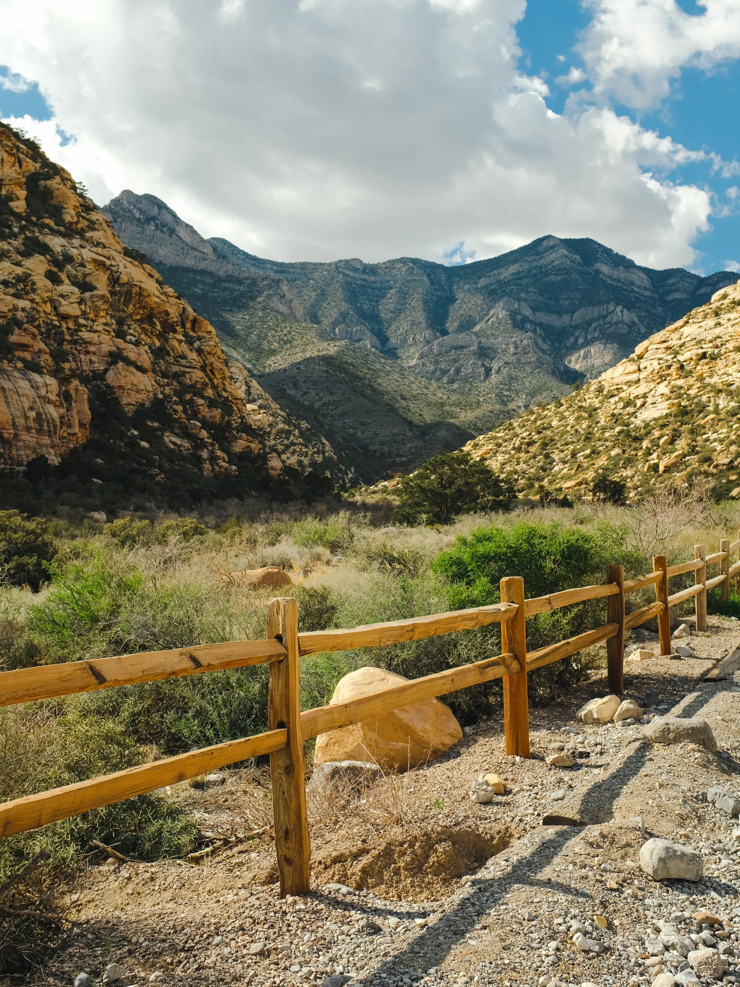 A scenic mountain landscape with rocky hillsides, sparse vegetation, and a partly cloudy sky. A wooden fence runs along a dirt path in the foreground.