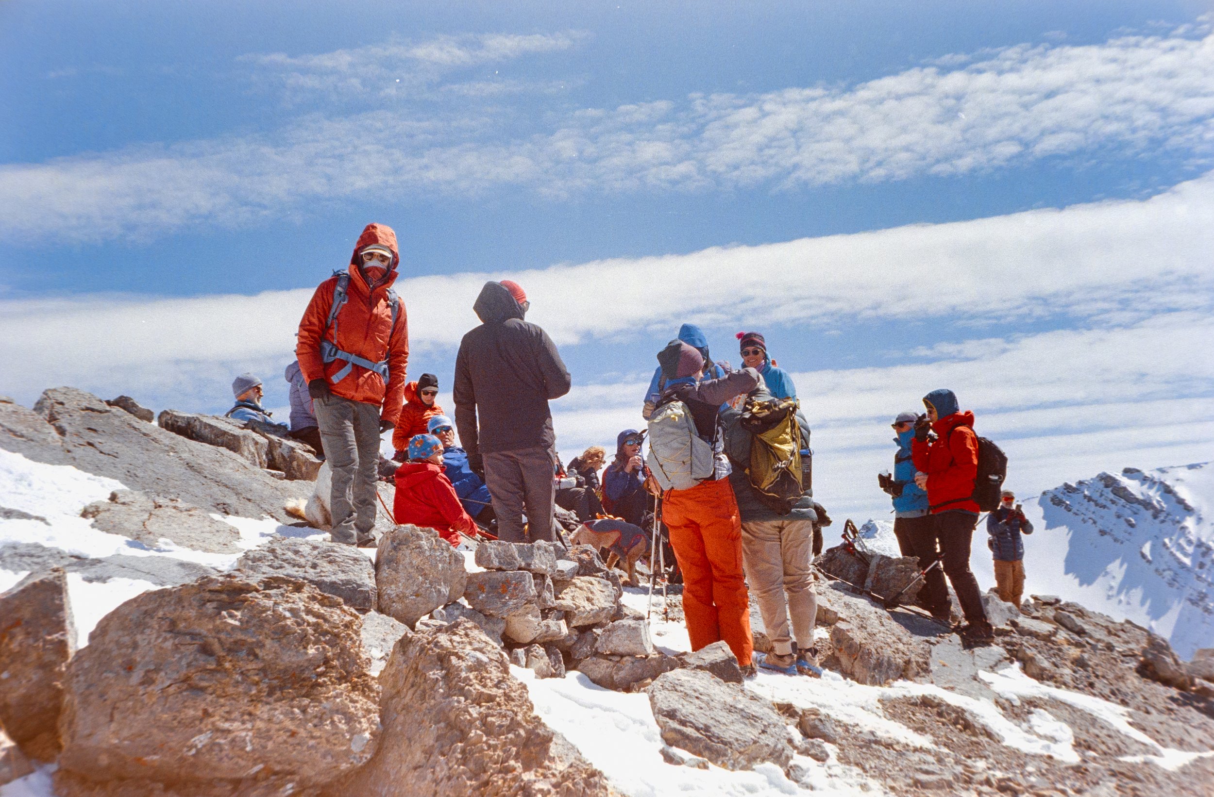 Group of hikers on a snowy mountain, some seated and some standing, wearing outdoor gear and backpacks, against a backdrop of snow-capped peaks and a partly cloudy sky.