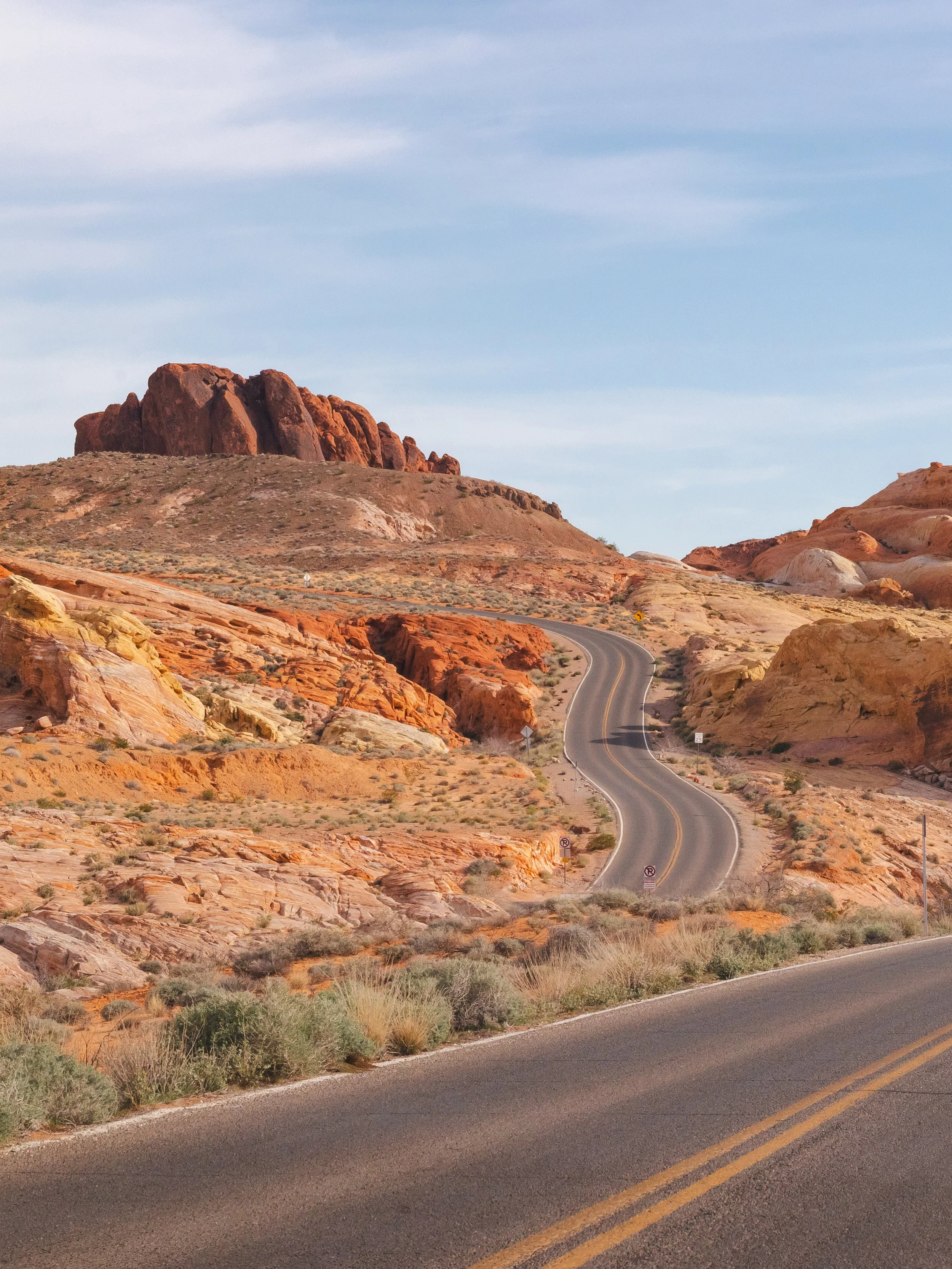 A winding asphalt road through a desert landscape with colorful rocks and large red rock formations under a cloudy sky.
