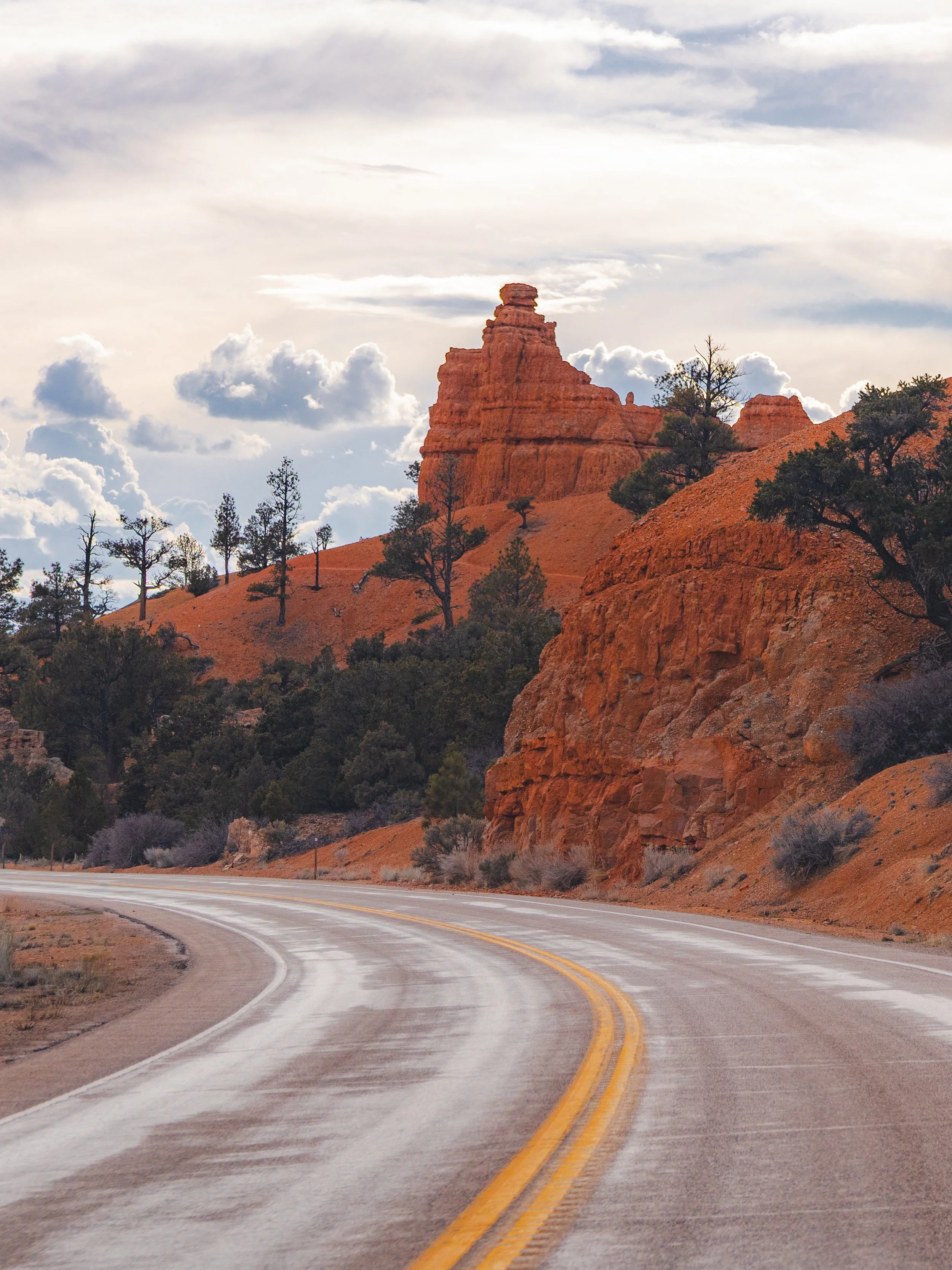 Winding highway passing through a desert landscape with orange rock formations and sparse trees, under a cloudy sky.