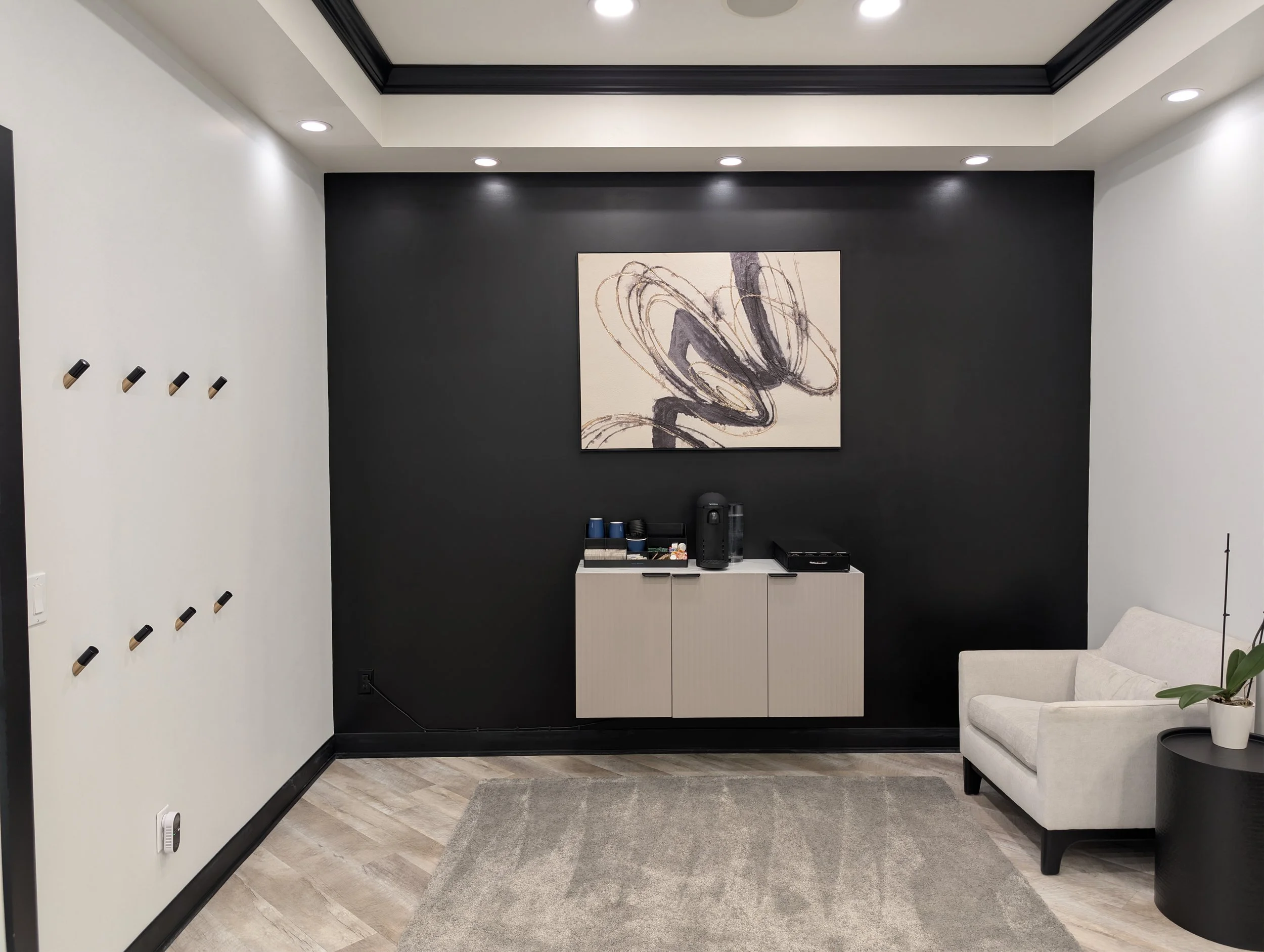 Modern living room corner with white armchair, black side table with plant, beige cabinet, black accent wall with abstract art, and coffee machine on the cabinet.