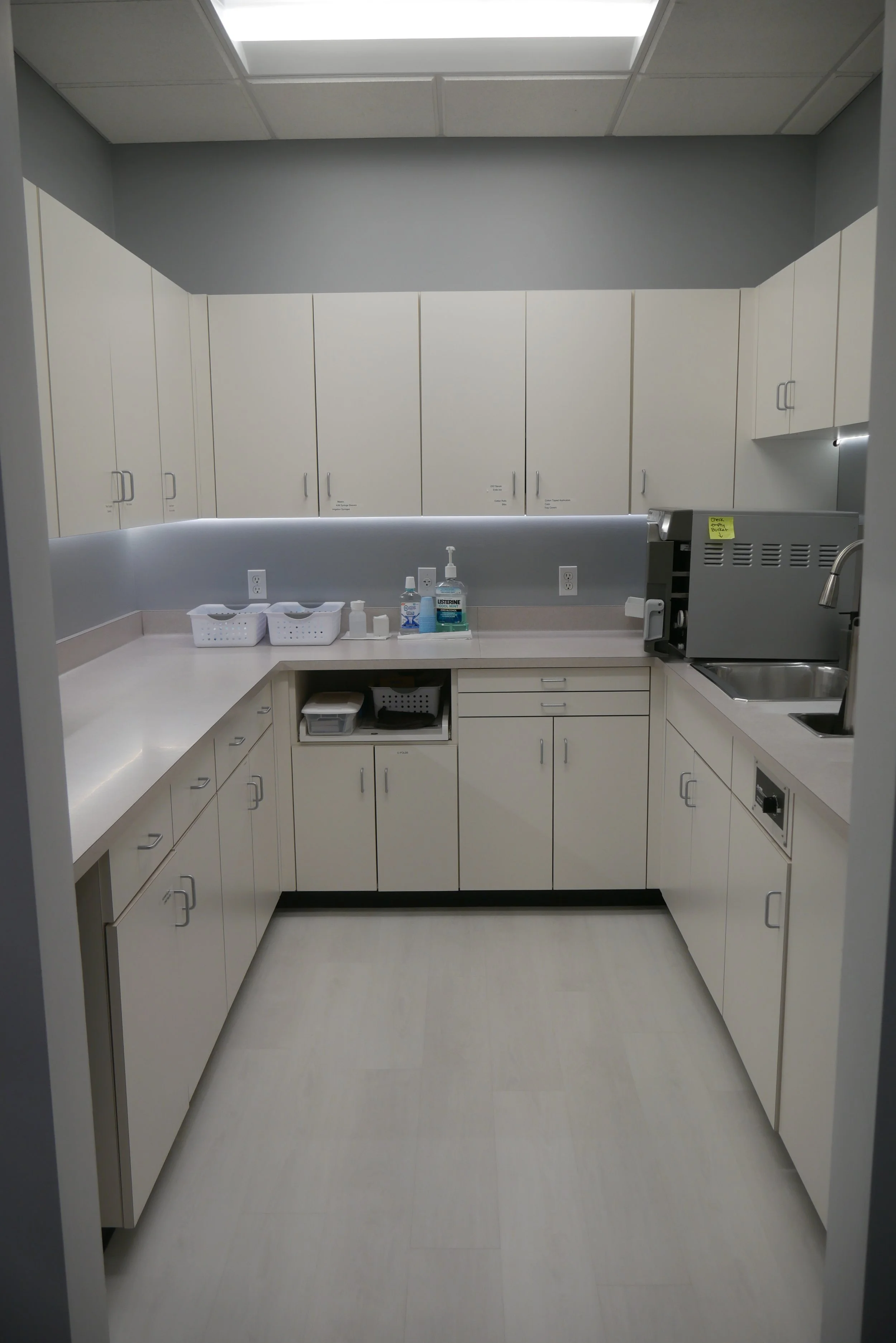 Empty medical office kitchen with white cabinets, countertop essentials, and a small sink.