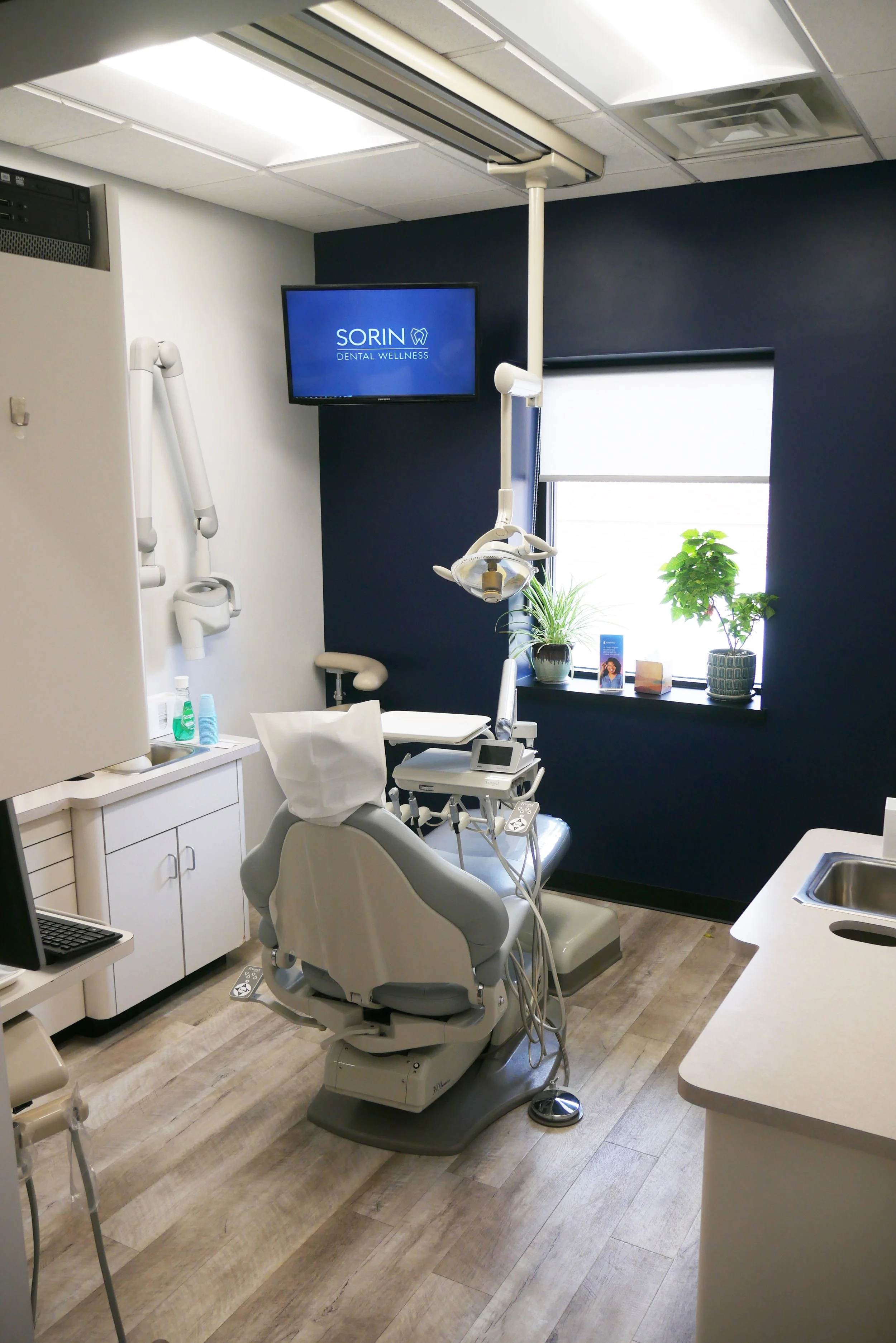 A dental examination room with a dental chair, overhead dental light, television screen, and a window with potted plants.