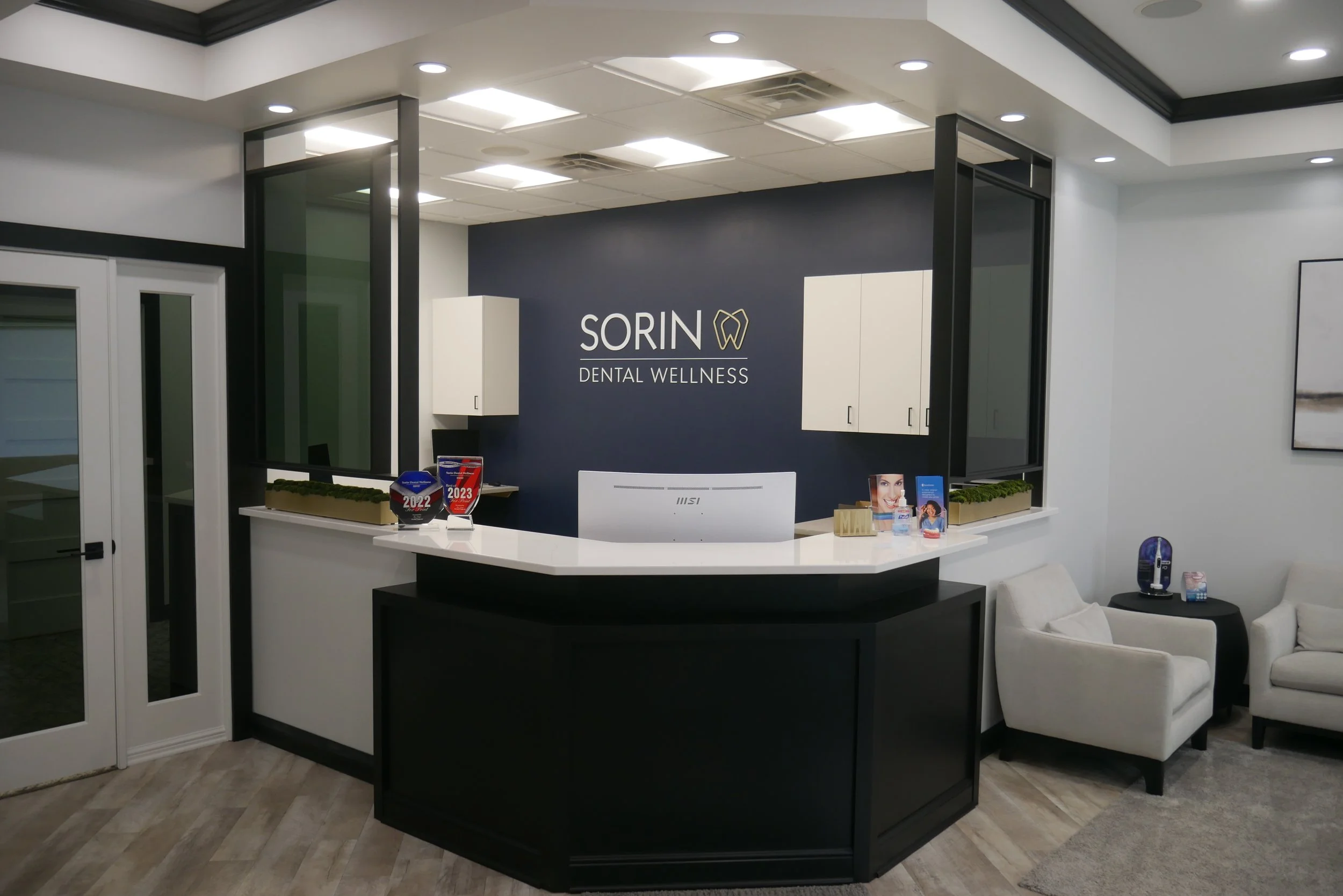 Reception desk at Sorin Dental Wellness with computer, awards, brochures, and seating area with chair and armchair.