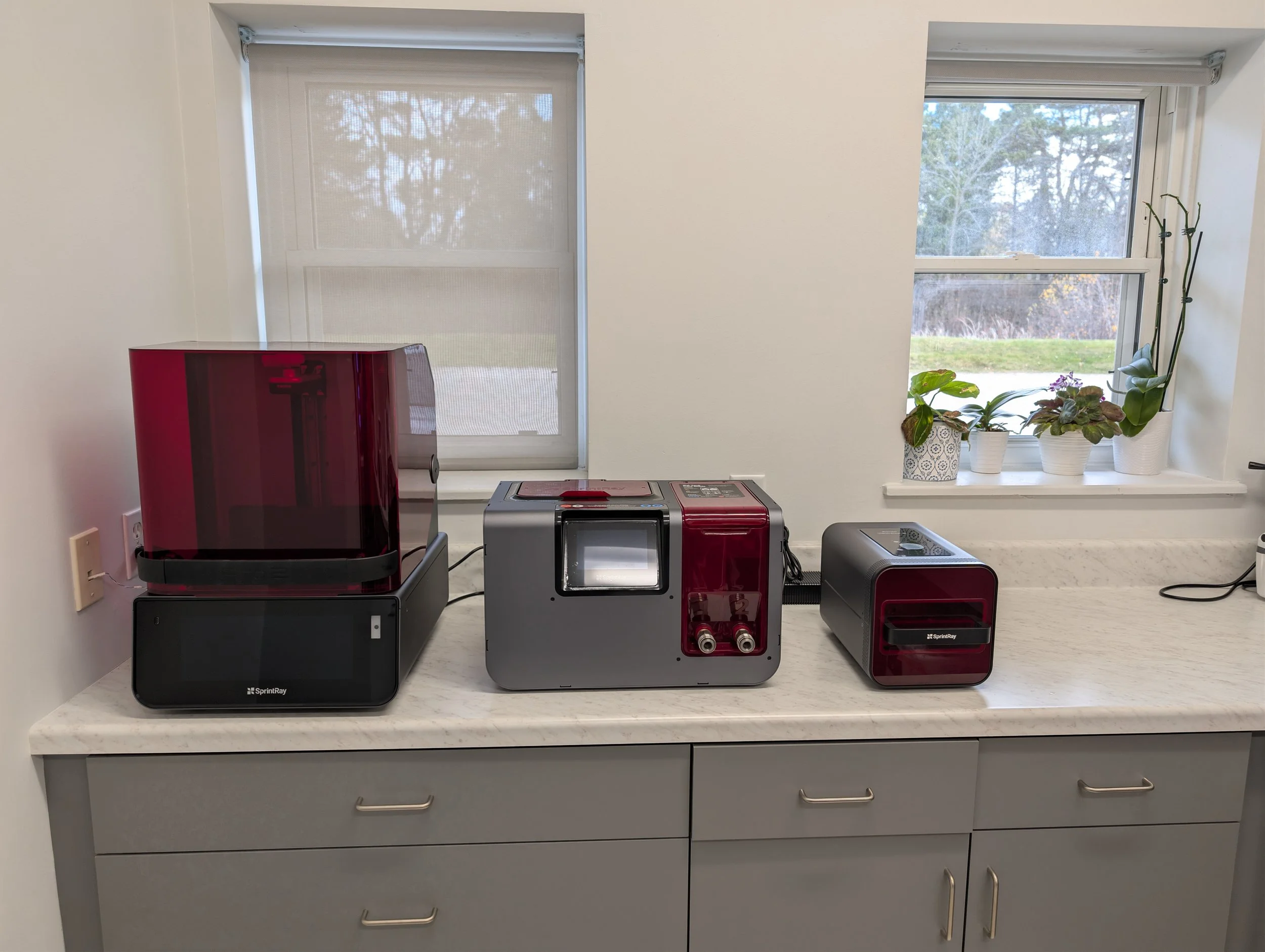 Laboratory setup with multiple scientific instruments on a white countertop, with windows in the background showing an outdoor scene with trees and plants.