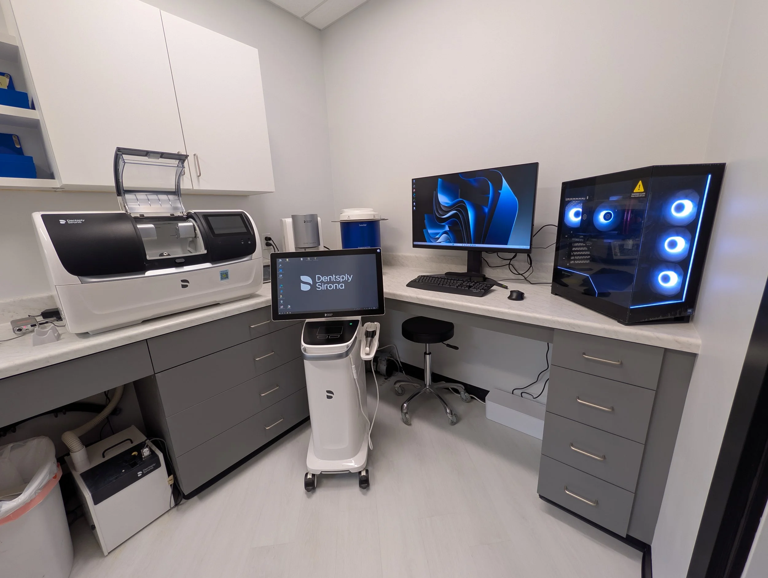 Dental office workspace with a computer, large monitor, 3D printer, and dental scanner.