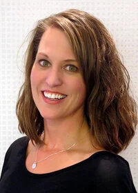 Portrait of a smiling woman with shoulder-length wavy brown hair, green eyes, wearing a black top and silver necklace, against a white background.