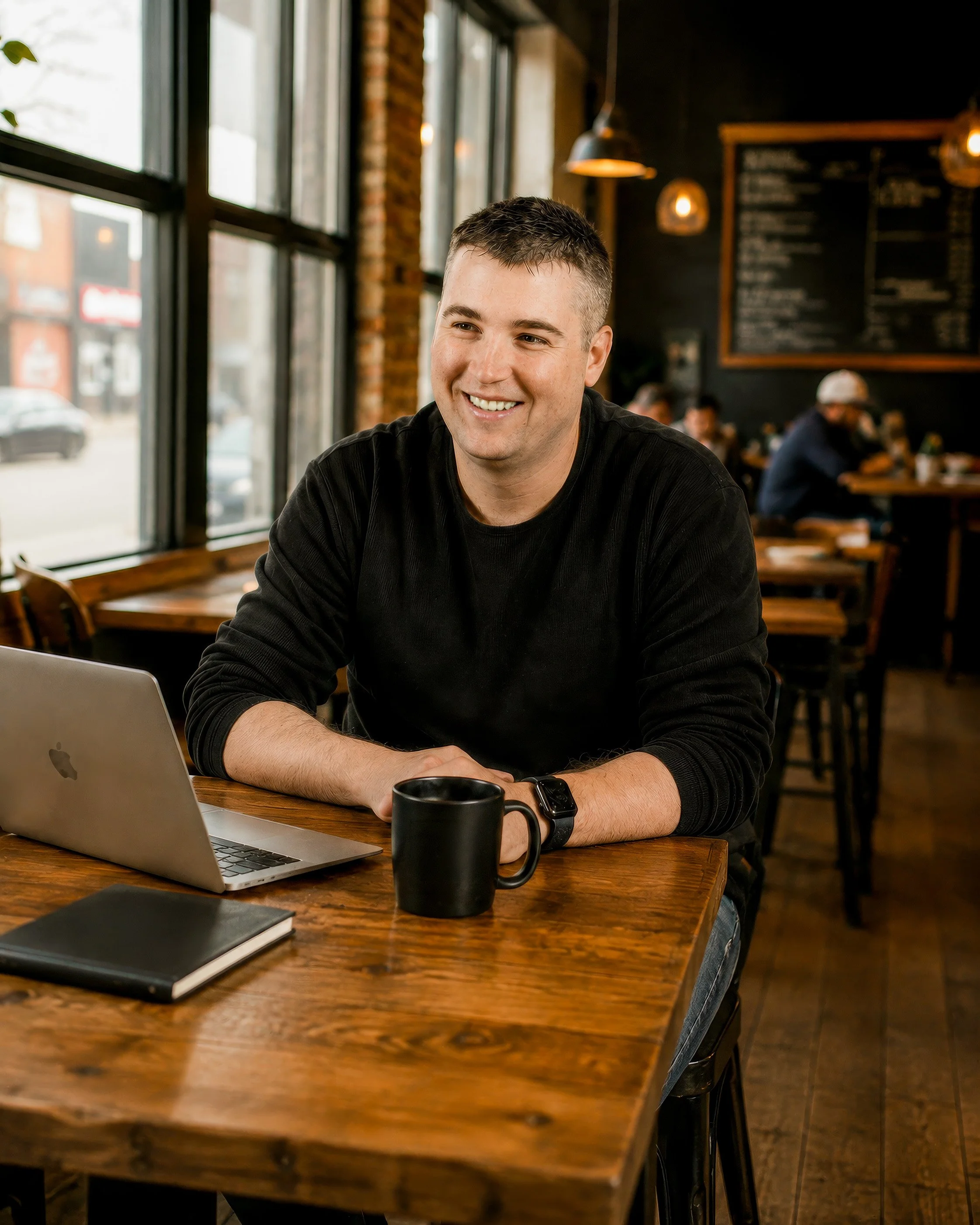 A smiling man sitting at a wooden table in a coffee shop with a laptop, a black mug, and a closed notebook in front of him. The background shows other patrons and a black wall with a menu board, large windows on the left letting in natural light, and hanging lights.