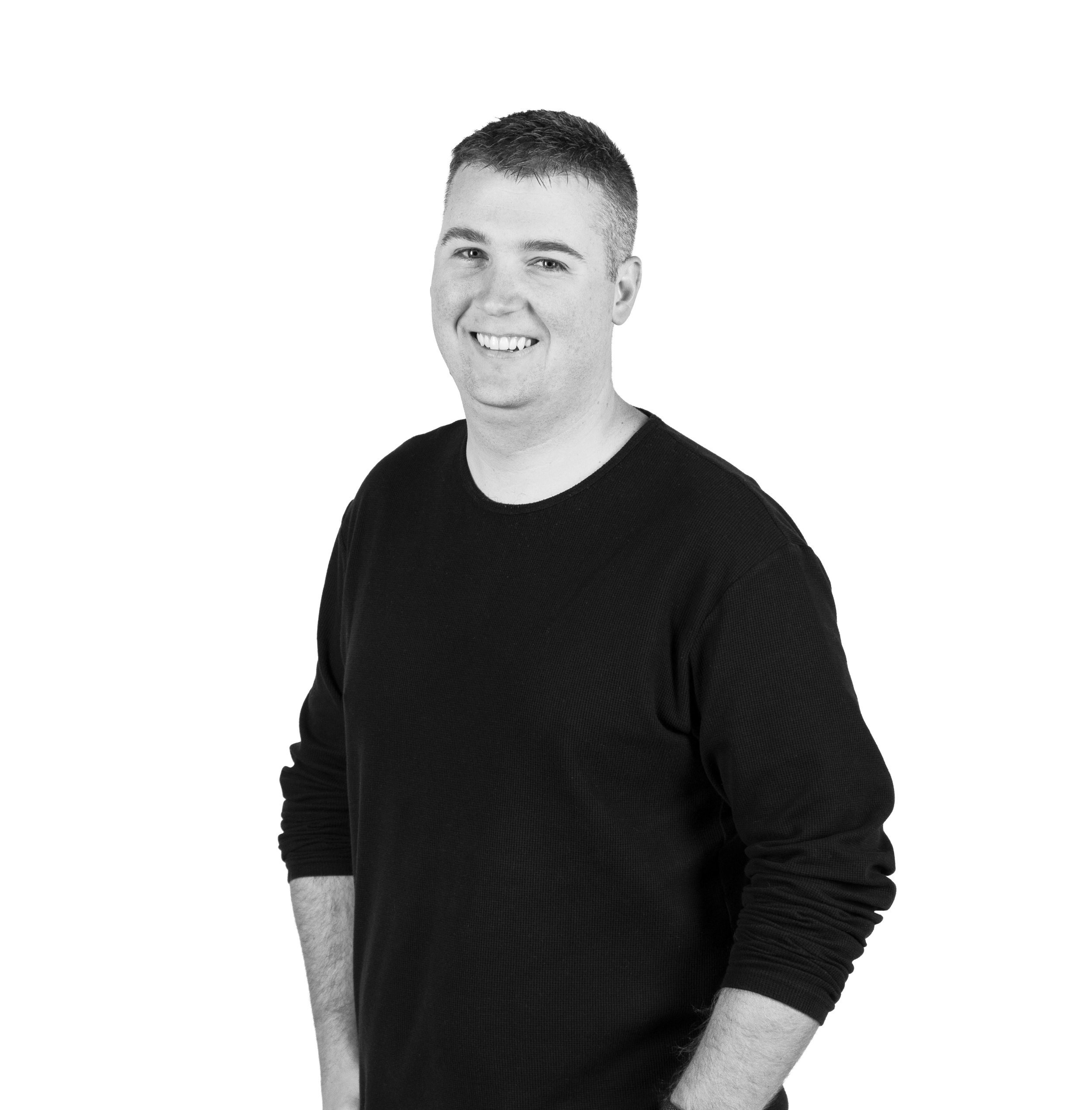 Black and white photo of a smiling young man with short hair, wearing a long-sleeve black shirt, standing against a plain background.