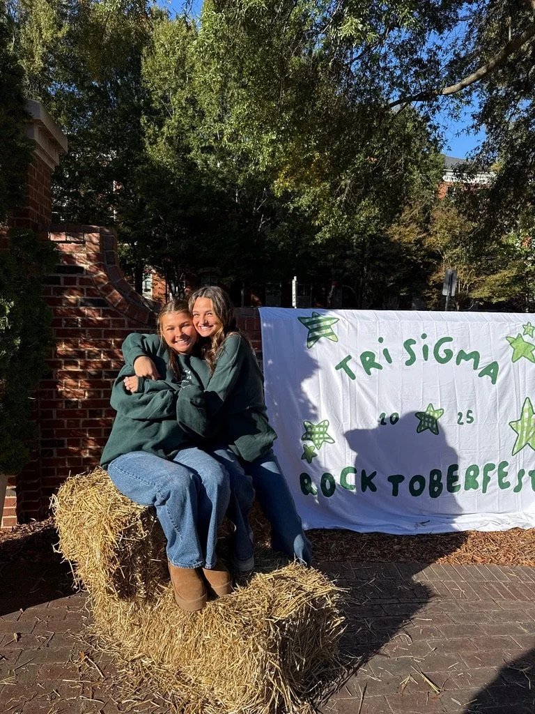 Two girls smiling and hugging while sitting on a hay bale in front of a white banner with holiday-themed decorations. The banner reads 'Trisigma Rocktoberfest 2025' with green stars.