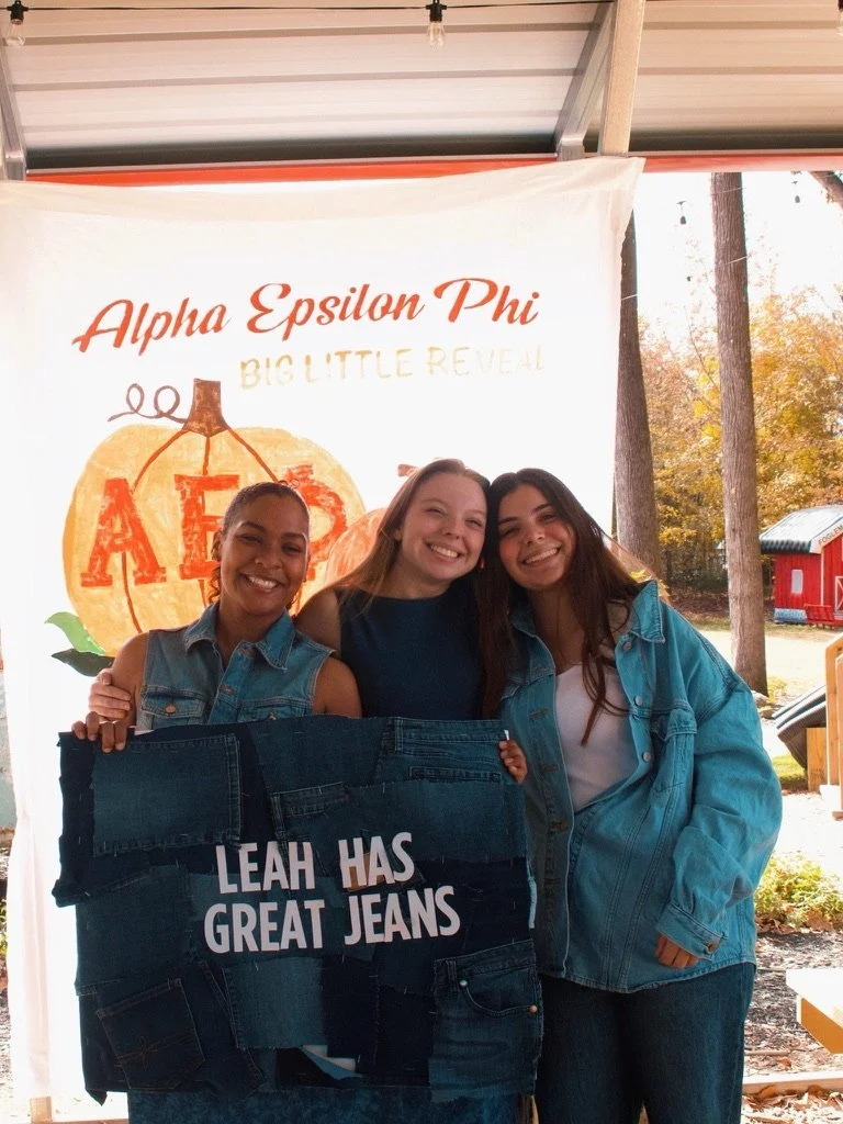 Three women standing outdoors, smiling, holding a sign that says "Leah has great jeans," with a background banner reading "Alpha Epsilon Phi Big Little Reveal" and fall trees.