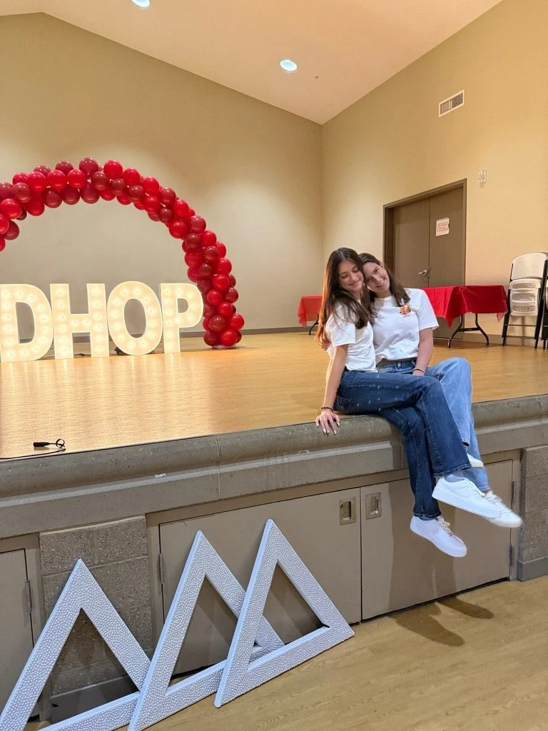 Two young women sitting on the edge of a stage with a backdrop of red balloon arch and large illuminated letters spelling "DHOP." The stage has red tablecloths and stacked chairs. 