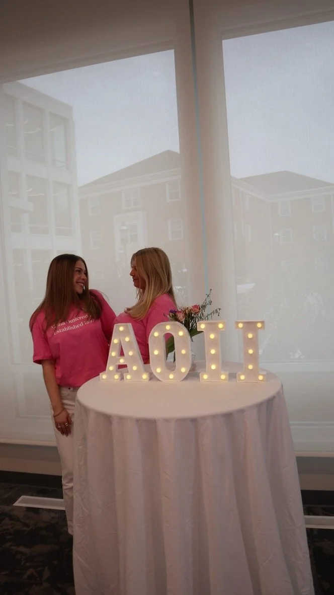 Two women in pink shirts smiling and looking at each other near a round table with illuminated letters spelling out 'AOII' and a small floral arrangement, in front of a large window.