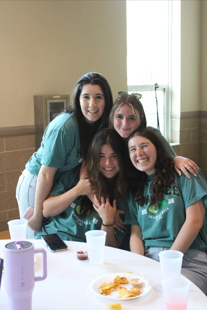 Four young women smiling and hugging around a table at an indoor gathering. 