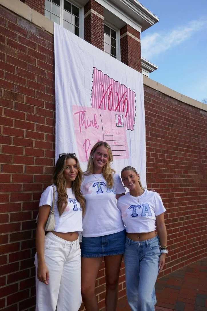 Three young women wearing white crop-top T-shirts with Greek letters (standing in front of a brick wall with a large pink-and-white breast cancer awareness banner and a red ribbon symbol, under a blue sky).