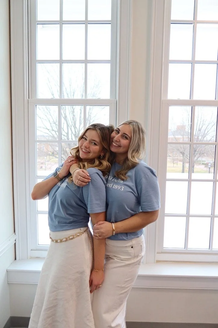 Two women smiling and hugging standing in front of large windows inside a building.