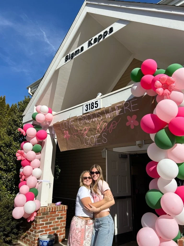 Two women with sunglasses hugging in front of a decorated house entrance with pink, white, and green balloons and a sign that reads "Love grows where Sig Kap goes".