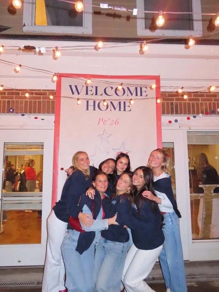 Group of six women smiling and hugging in front of a welcome home sign at a gathering, with string lights overhead.