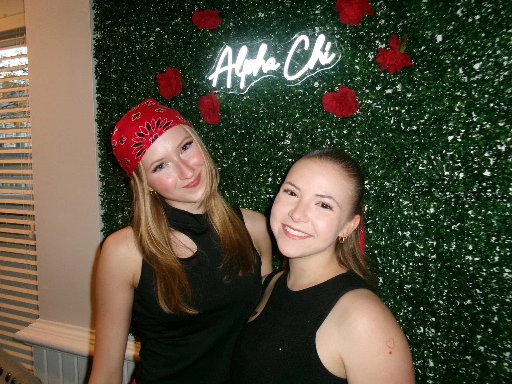 Two women smiling and posing in front of a green wall with red decorations and a neon sign that says "Alpha Chi."