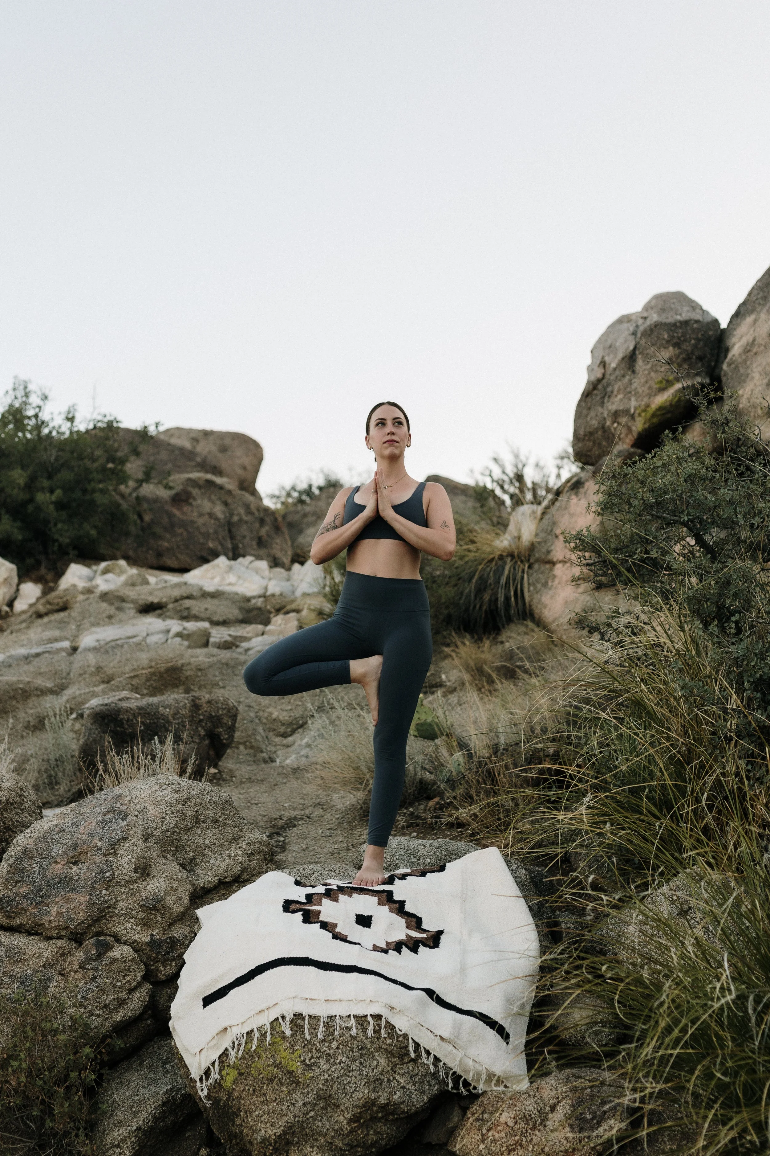 A woman practicing yoga outdoors on a rocky terrain, standing on a patterned rug, balancing in a tree pose with hands in prayer position.