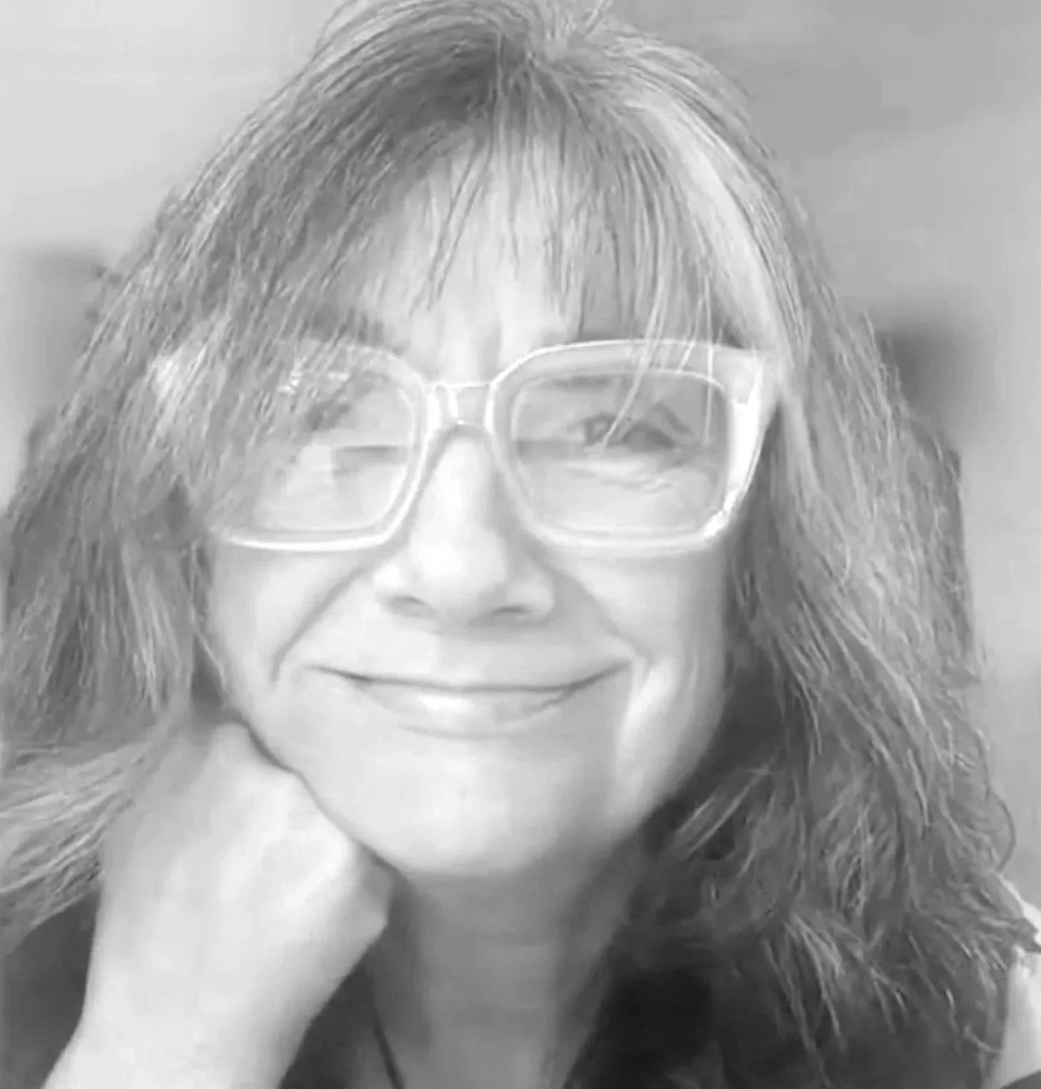 A smiling woman with long gray hair, wearing a black jacket and a heart-shaped pendant necklace, outdoors in front of a blurred natural background.