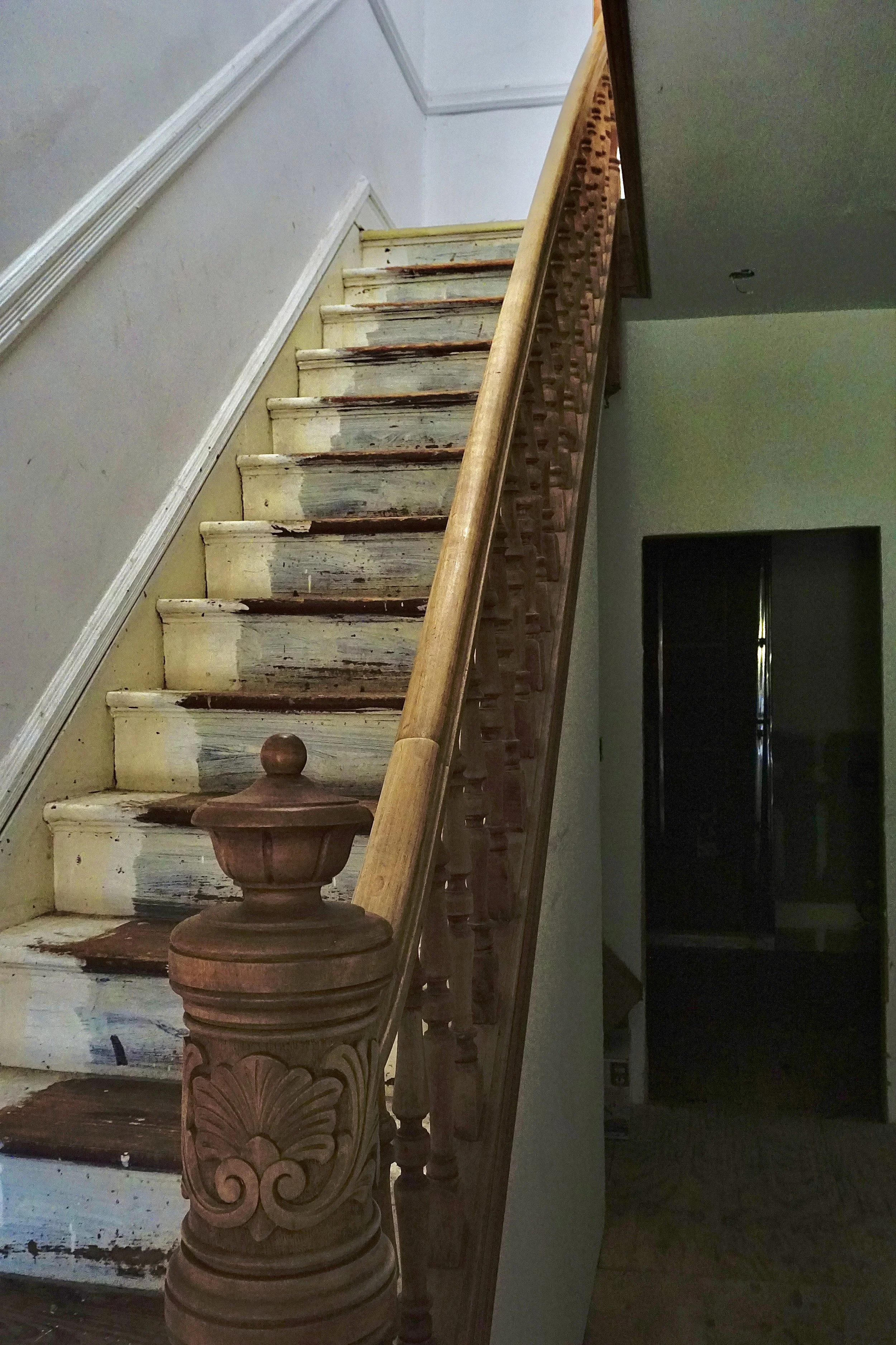 An old wooden staircase with peeling white paint and a carved wooden newel post at the bottom. The stairs lead to an upper floor, with some parts showing exposed wood and signs of wear.