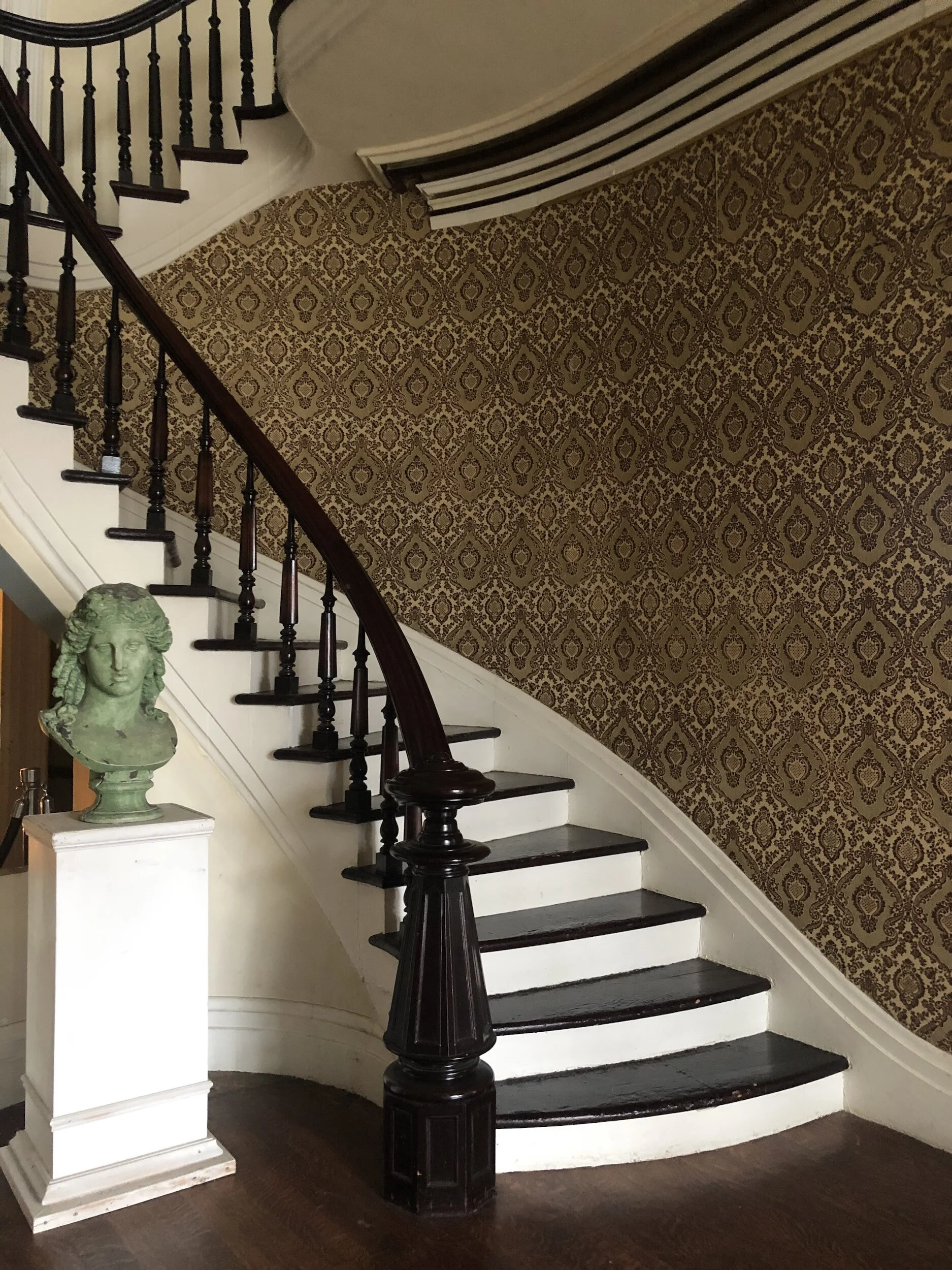 Indoor curved staircase with dark wooden handrail and white risers, wallpaper with gold and reddish-brown pattern on the wall, and a green bust sculpture on a white pedestal.